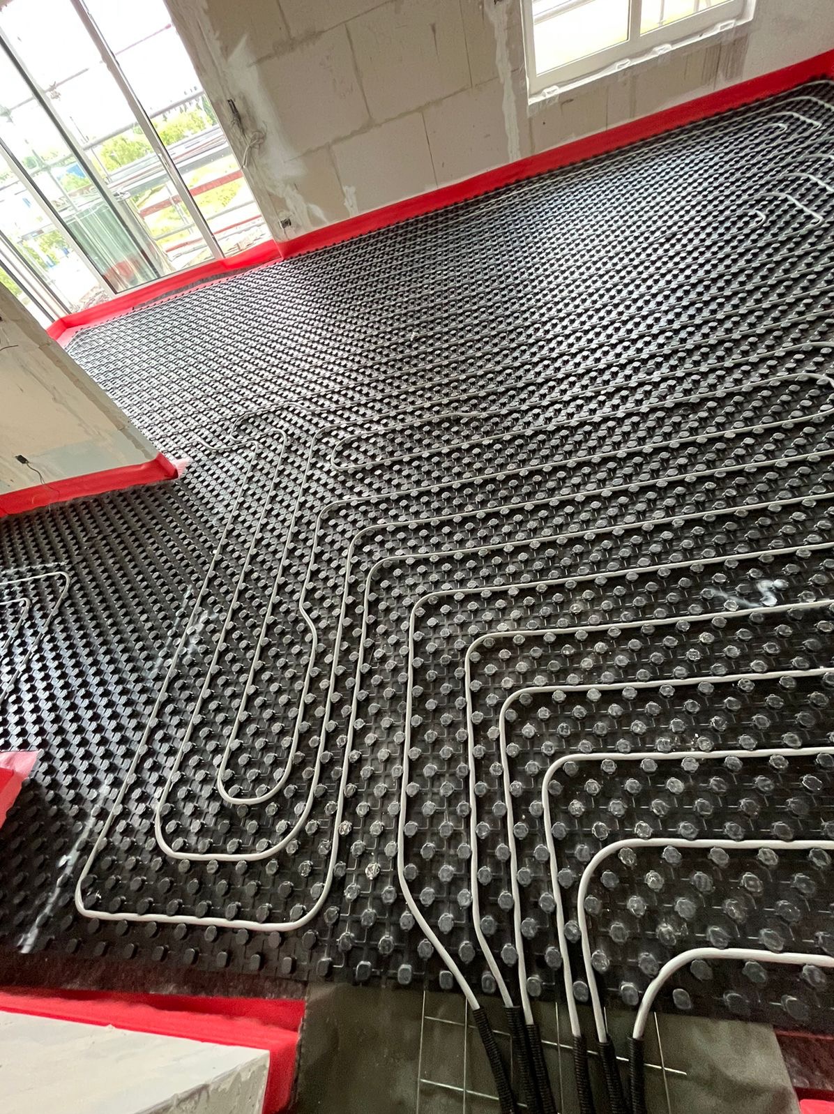 Rozłożona instalacja ogrzewania podłogowego na czarnej macie z wypustkami, widoczne białe rurki w kształcie pętli i fragmenty czerwonej taśmy na obrzeżach, surowy stan wykończenia wnętrza.