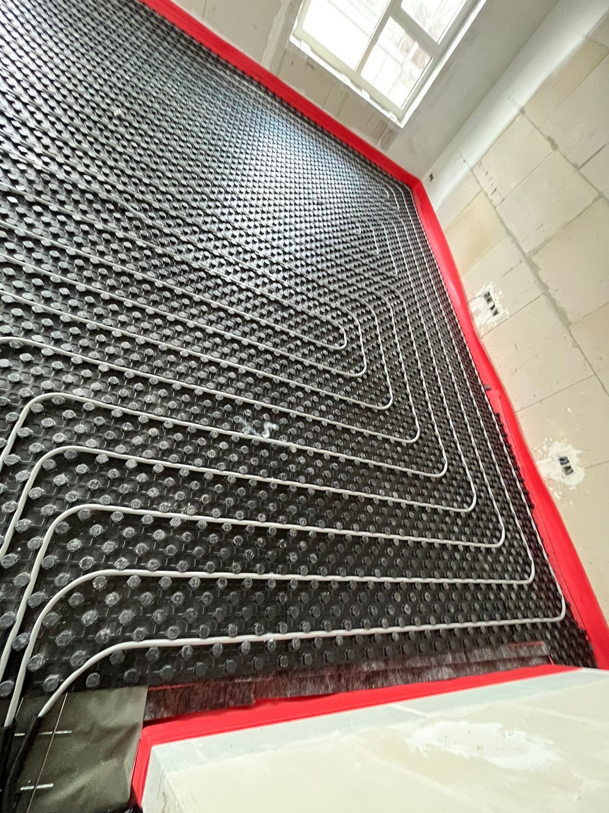 Czarna mata z wypustkami i białymi rurkami instalacji podłogowej, ułożona w surowym pomieszczeniu z oknem, zabezpieczona czerwoną taśmą.