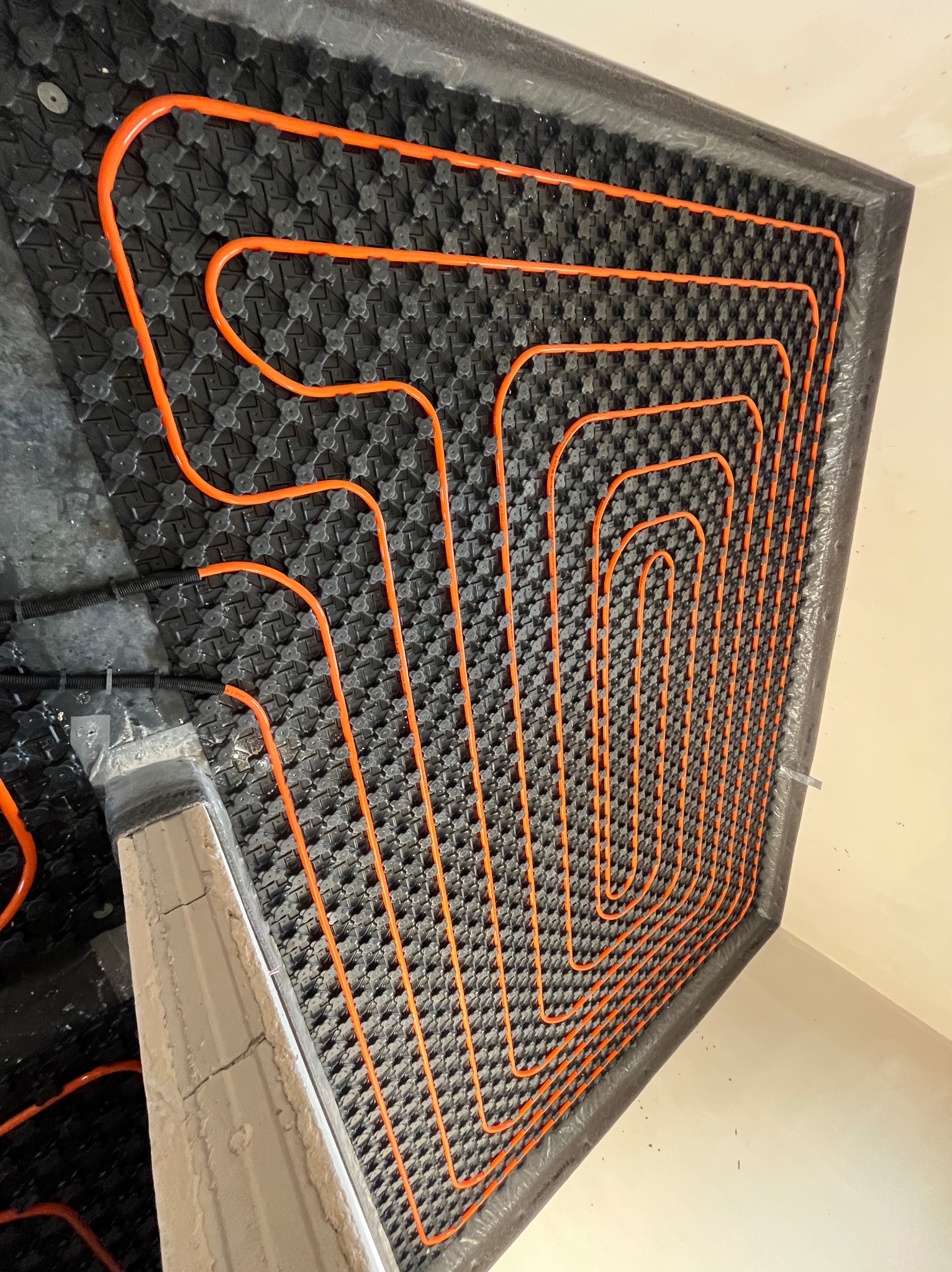 Pomarańczowe rurki układane w pętli na czarnej, perforowanej macie pod system ogrzewania podłogowego, widoczne fragmenty wylewki i instalacji elektrycznej.