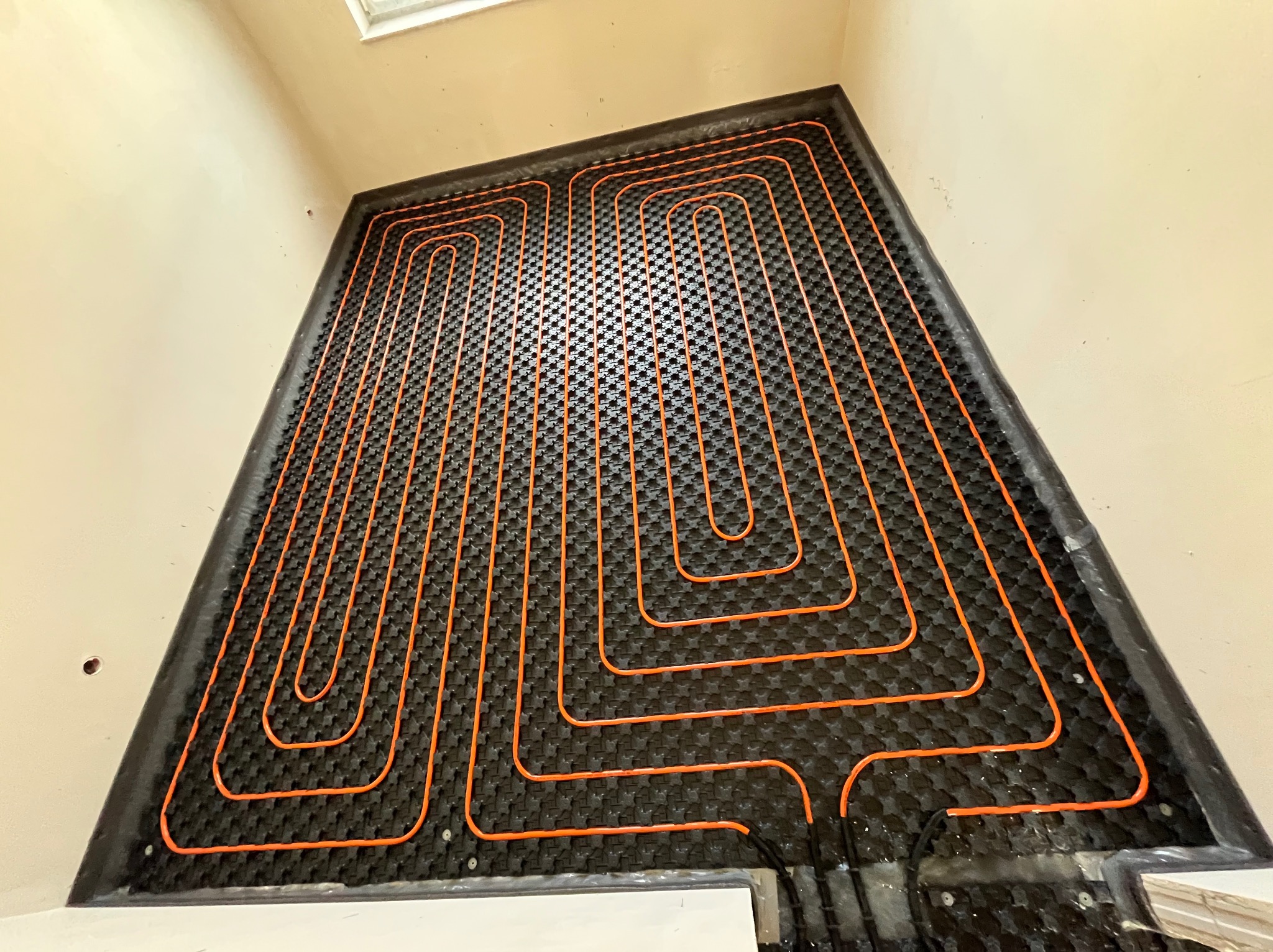 Pomarańczowe rury ułożone w pętle na czarnej macie grzewczej w trakcie instalacji systemu podłogowego, widok z góry, przy ścianie z widocznymi przewodami.