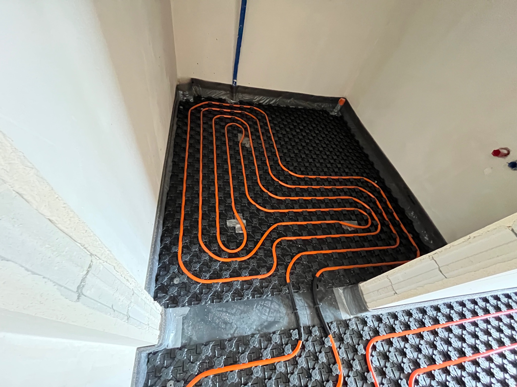 Pomarańczowe rury ułożone na czarnej macie w systemie ogrzewania podłogowego, widoczne fragmenty ścian i narożnik pomieszczenia w trakcie montażu.