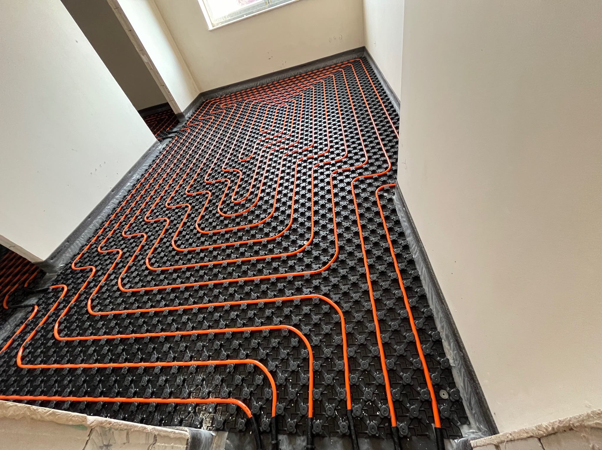 Montaż ogrzewania podłogowego: pomarańczowe rurki grzewcze ułożone na czarnej macie izolacyjnej w jasnym pomieszczeniu, widok z góry.
