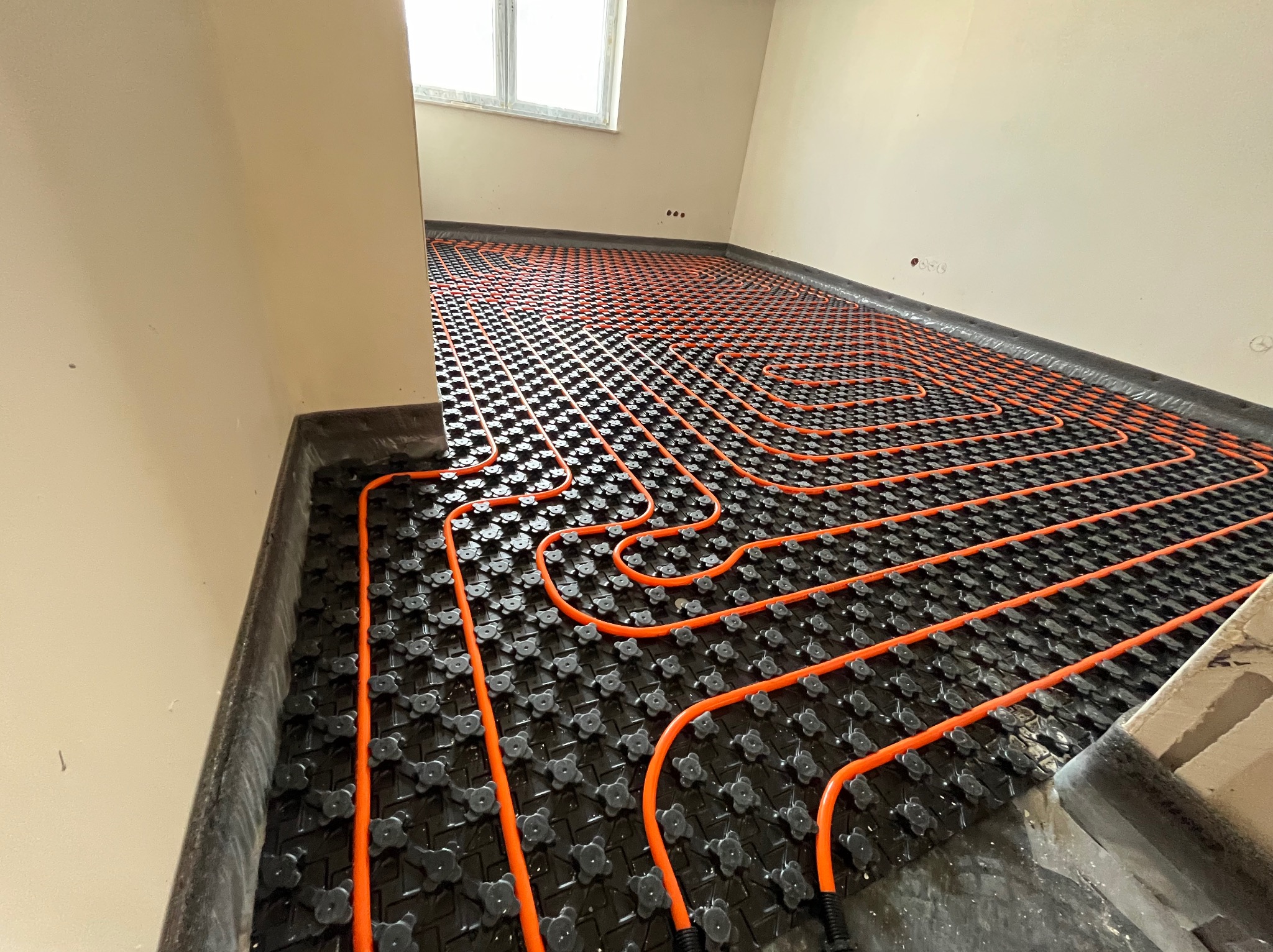 Pomarańczowe rury układane na czarnej macie z wypustkami, przygotowanie do wylewki pod ogrzewanie podłogowe w nowym budynku.