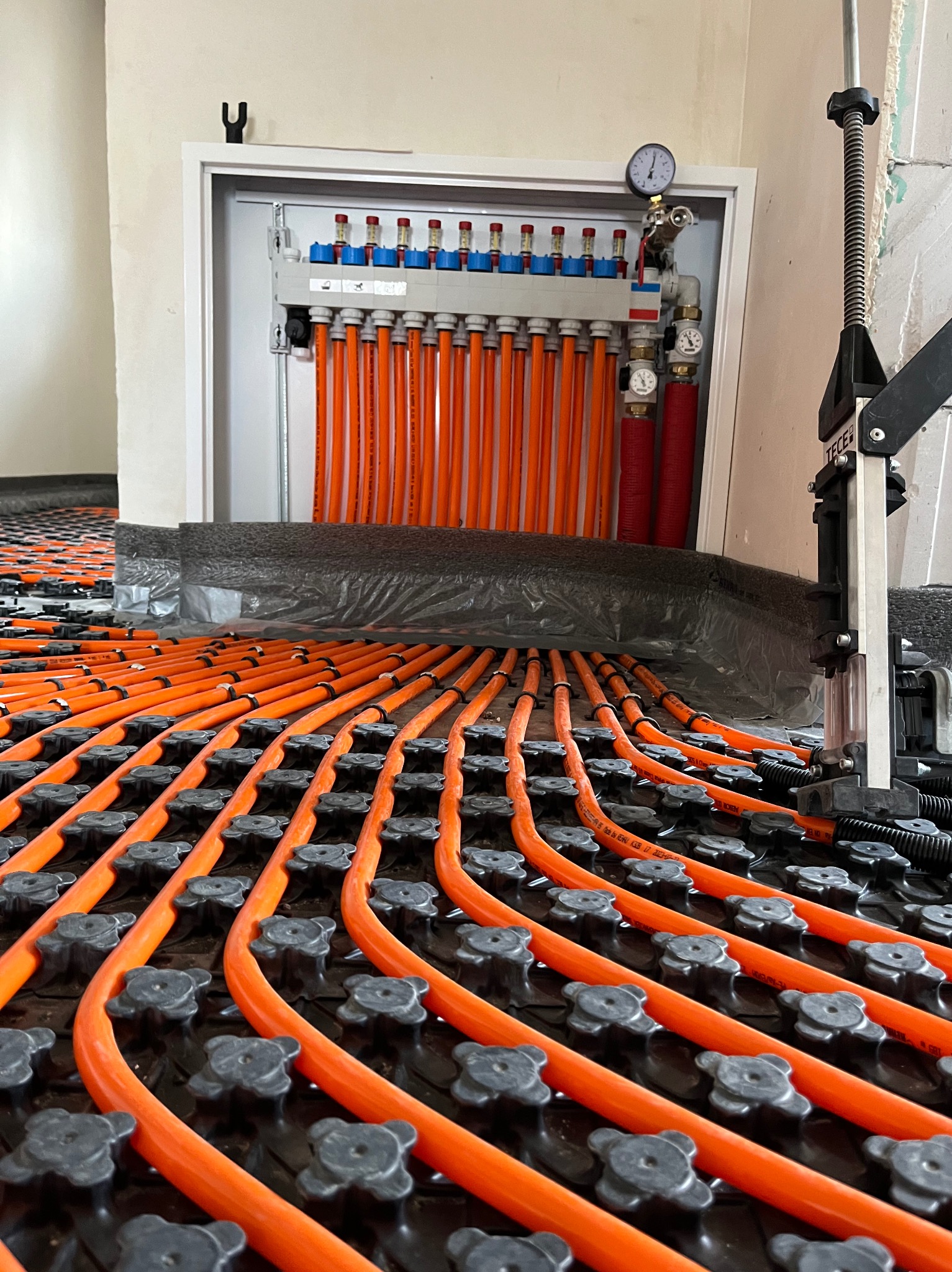 Pomarańczowe rury systemu ogrzewania podłogowego, zamocowane do czarnej maty, rozchodzące się od rozdzielacza w białej szafce, widoczne manometry i narzędzie do mocowania rur.