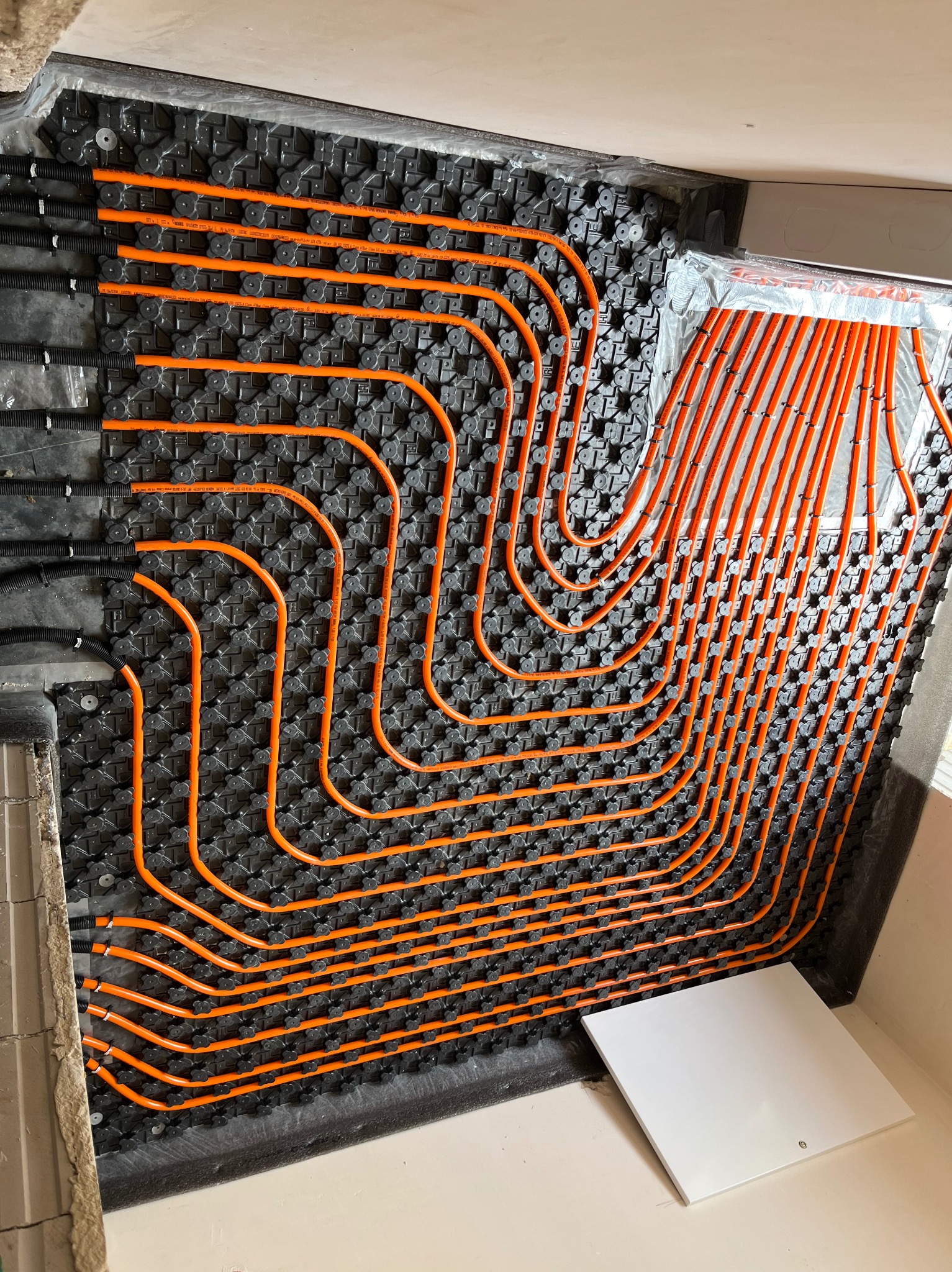 Pomarańczowe rury ułożone w pętle na czarnej macie montażowej, instalacja w systemie ogrzewania podłogowego, widoczny fragment ściany i biała płyta zakrywająca instalację.