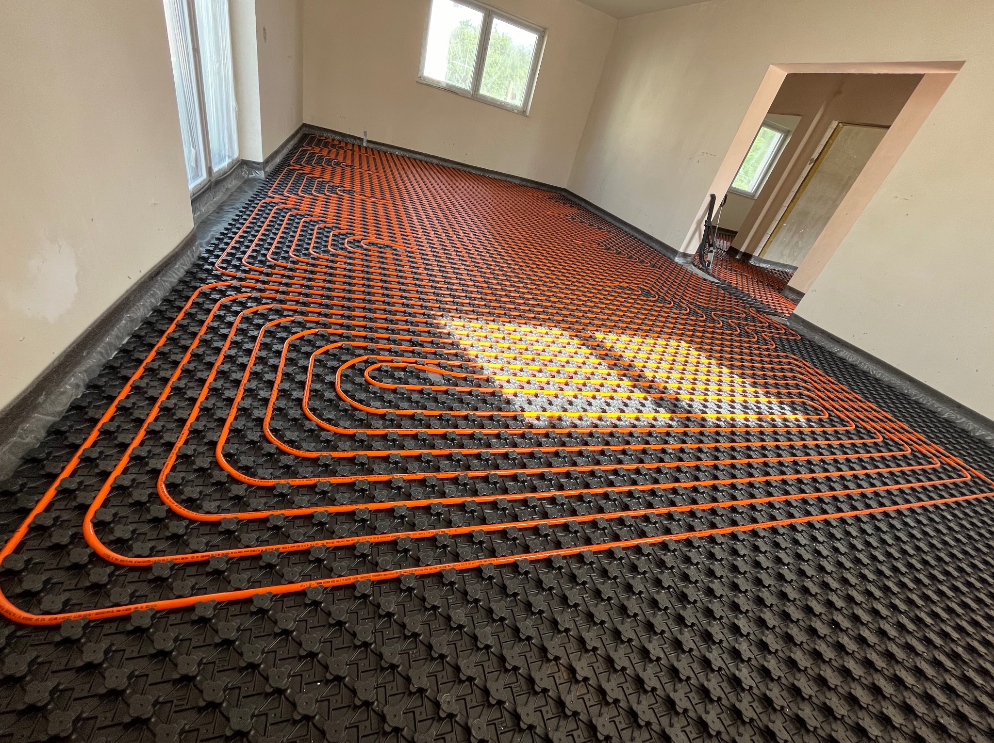 Pomarańczowe rury układane spiralnie na czarnej macie podłogowej, przygotowane do montażu ogrzewania podłogowego w jasnym pomieszczeniu z dwoma oknami.