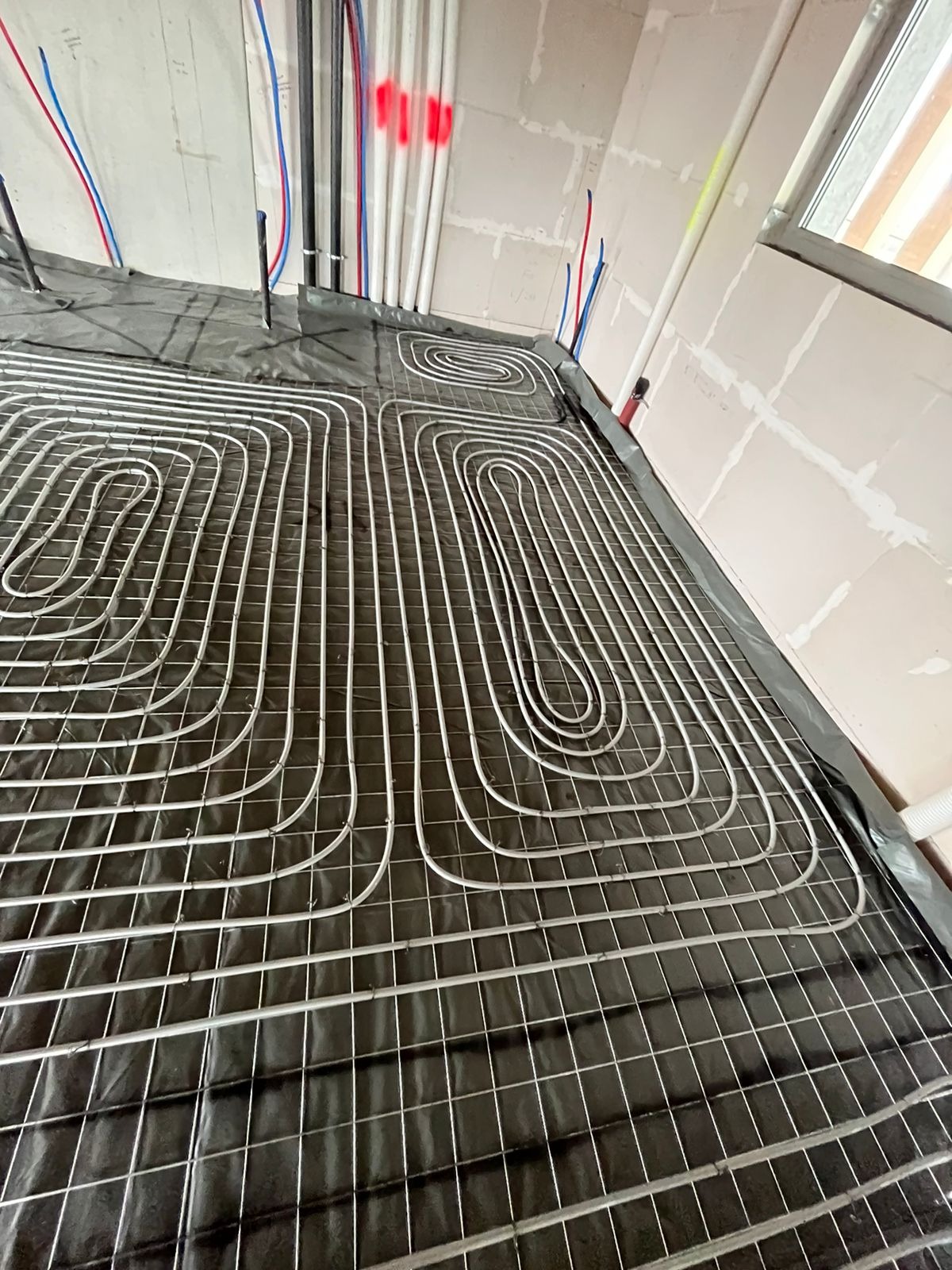 Montaż rur w systemie ogrzewania podłogowego na czarnej folii z siatką, widoczne rury doprowadzające na ścianie w surowym stanie.