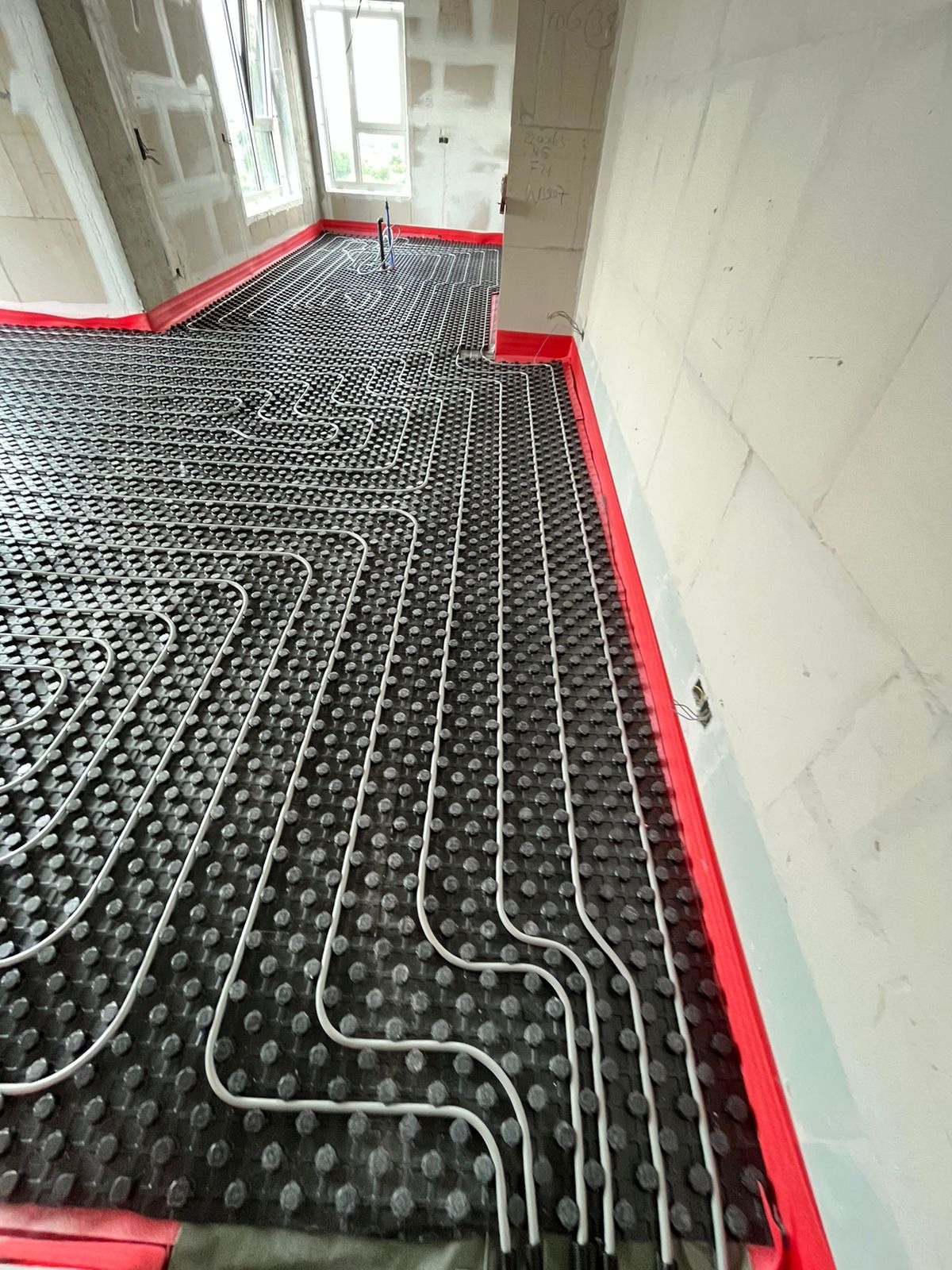 Rozłożona instalacja ogrzewania podłogowego na czarnej macie z wypustkami, zabezpieczona czerwoną taśmą przy ścianach w surowym wnętrzu z oknem i zapiskami na ścianie.