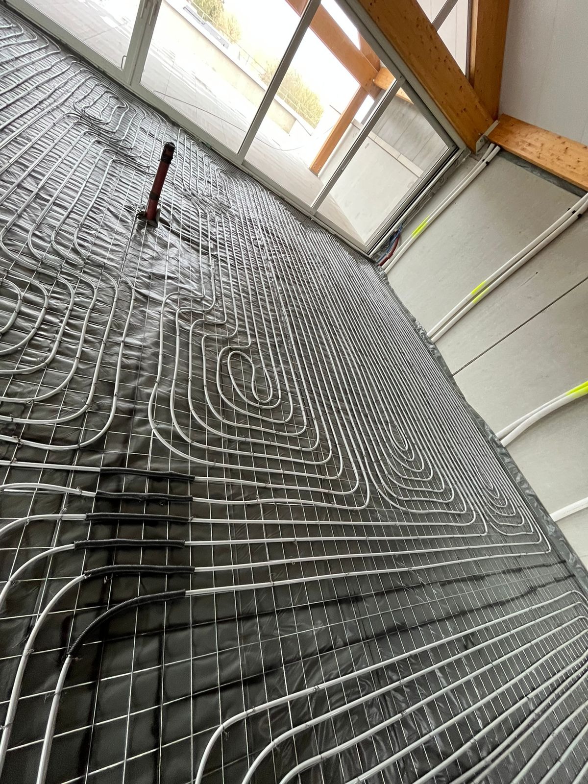 Rozłożone rury ogrzewania podłogowego na czarnej folii z siatką, widoczne okno i drewniana konstrukcja dachu