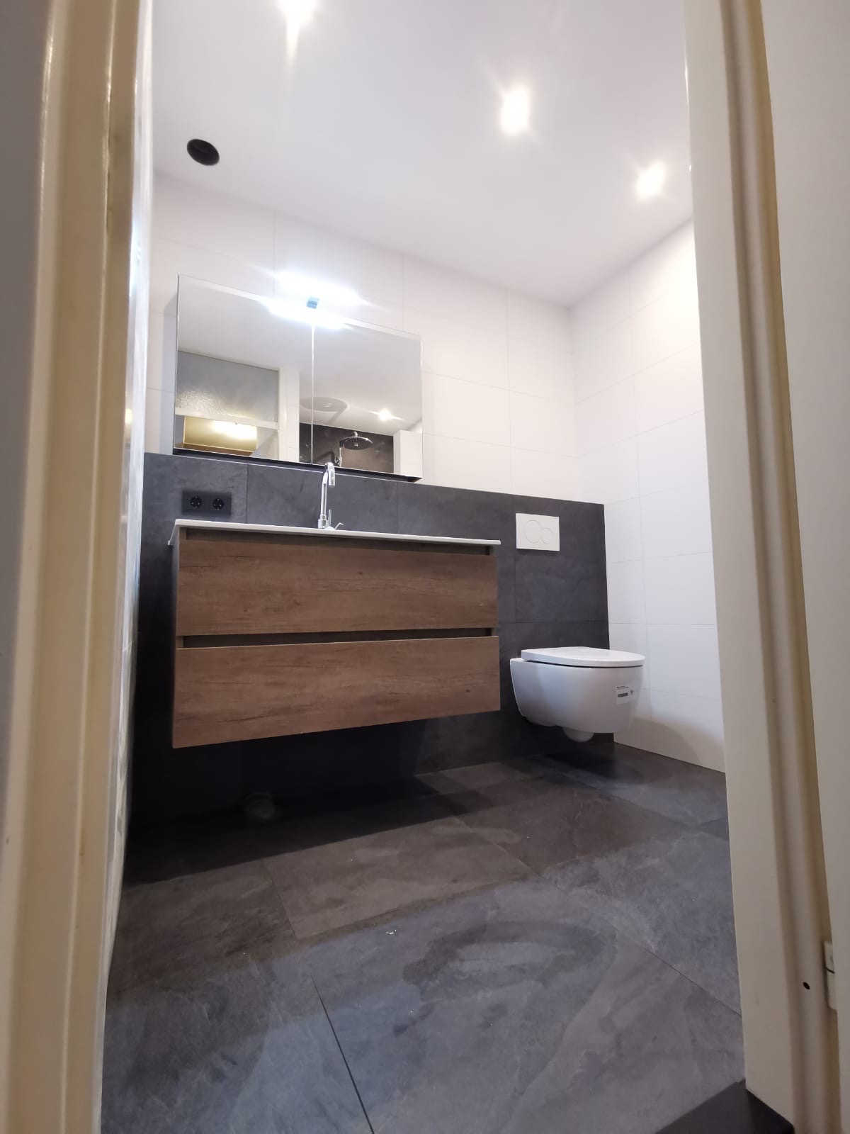 Nowoczesna łazienka z podwieszaną szafką z drewnopodobnymi frontami, umywalką, lustrem i podwieszaną toaletą, wykończona płytkami w odcieniach szarości i bieli, widok zza futryny drzwi.