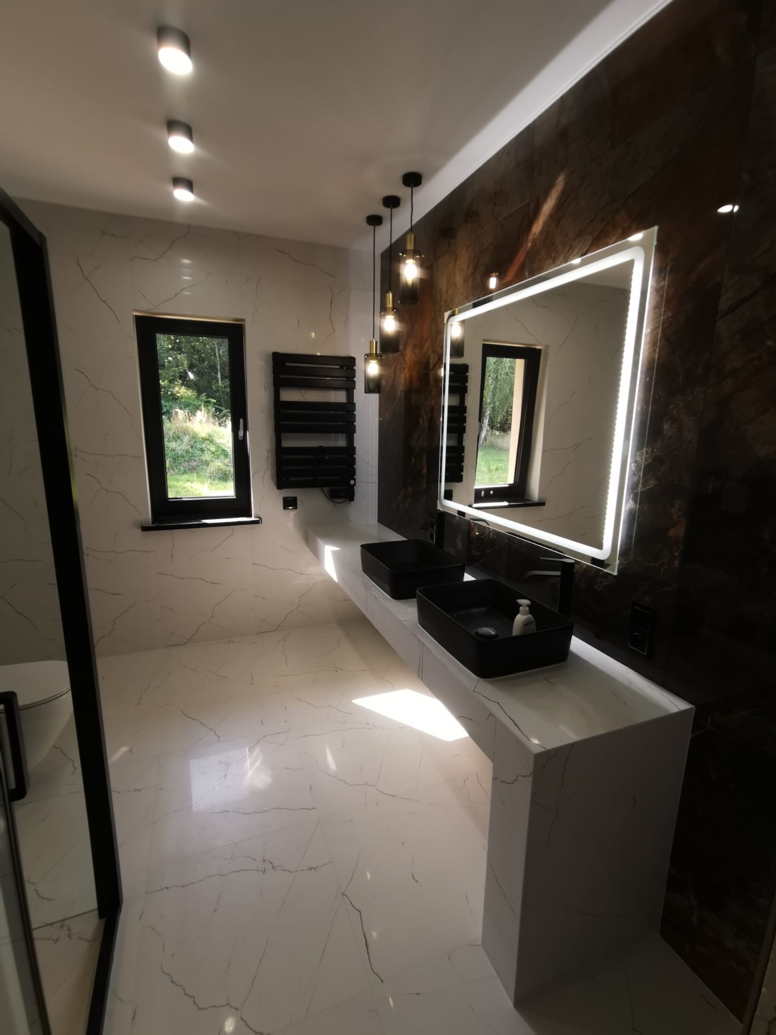 Nowoczesna łazienka z marmurową podłogą i ścianami, czarnymi umywalkami nablatowymi i podświetlanym lustrem LED, widok na okno z zielenią na zewnątrz.