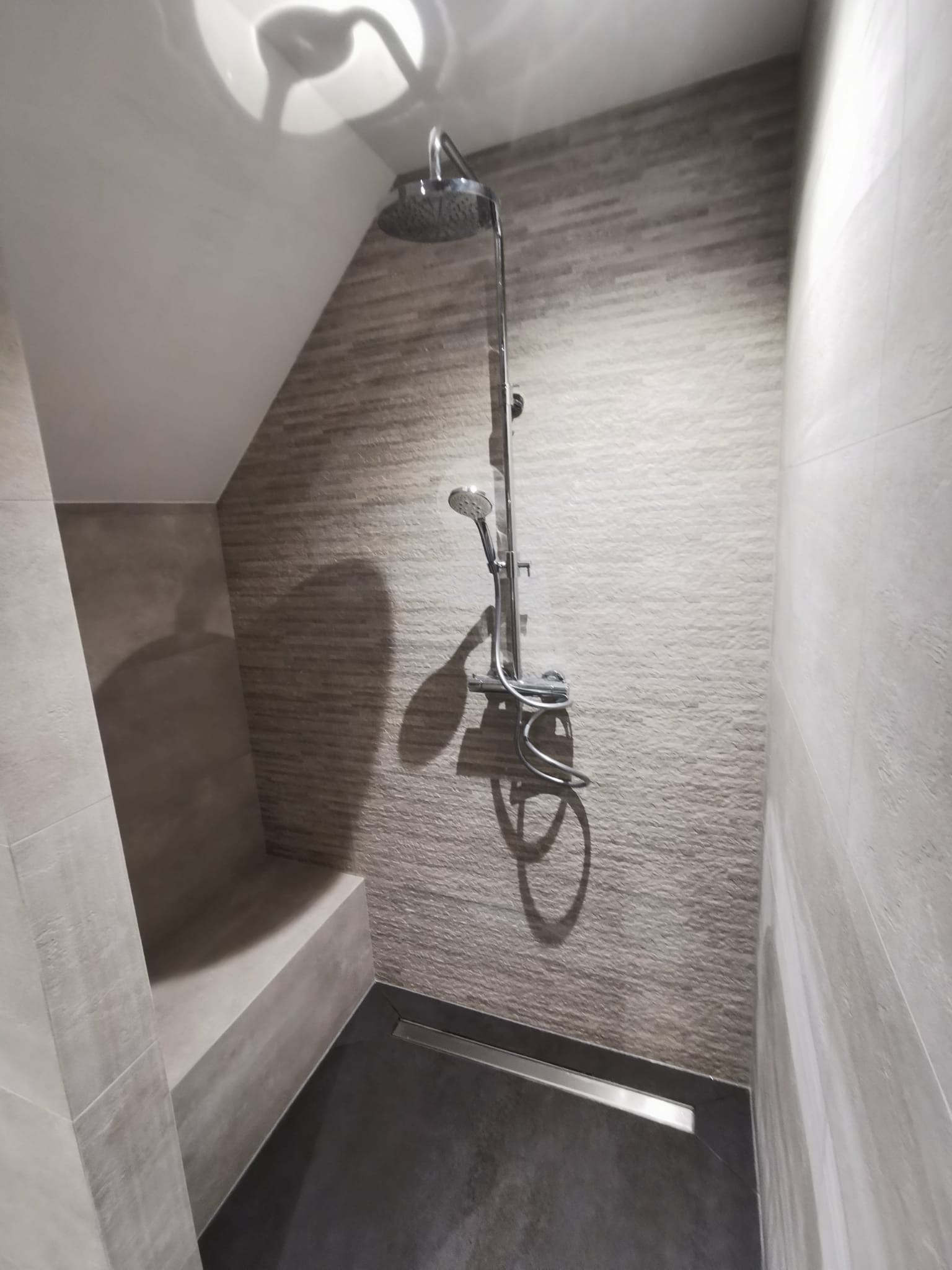 Wykończona kabina prysznicowa z siedziskiem, wyłożona szarymi płytkami imitującymi kamień w różnych odcieniach, z odpływem liniowym i zestawem natryskowym.