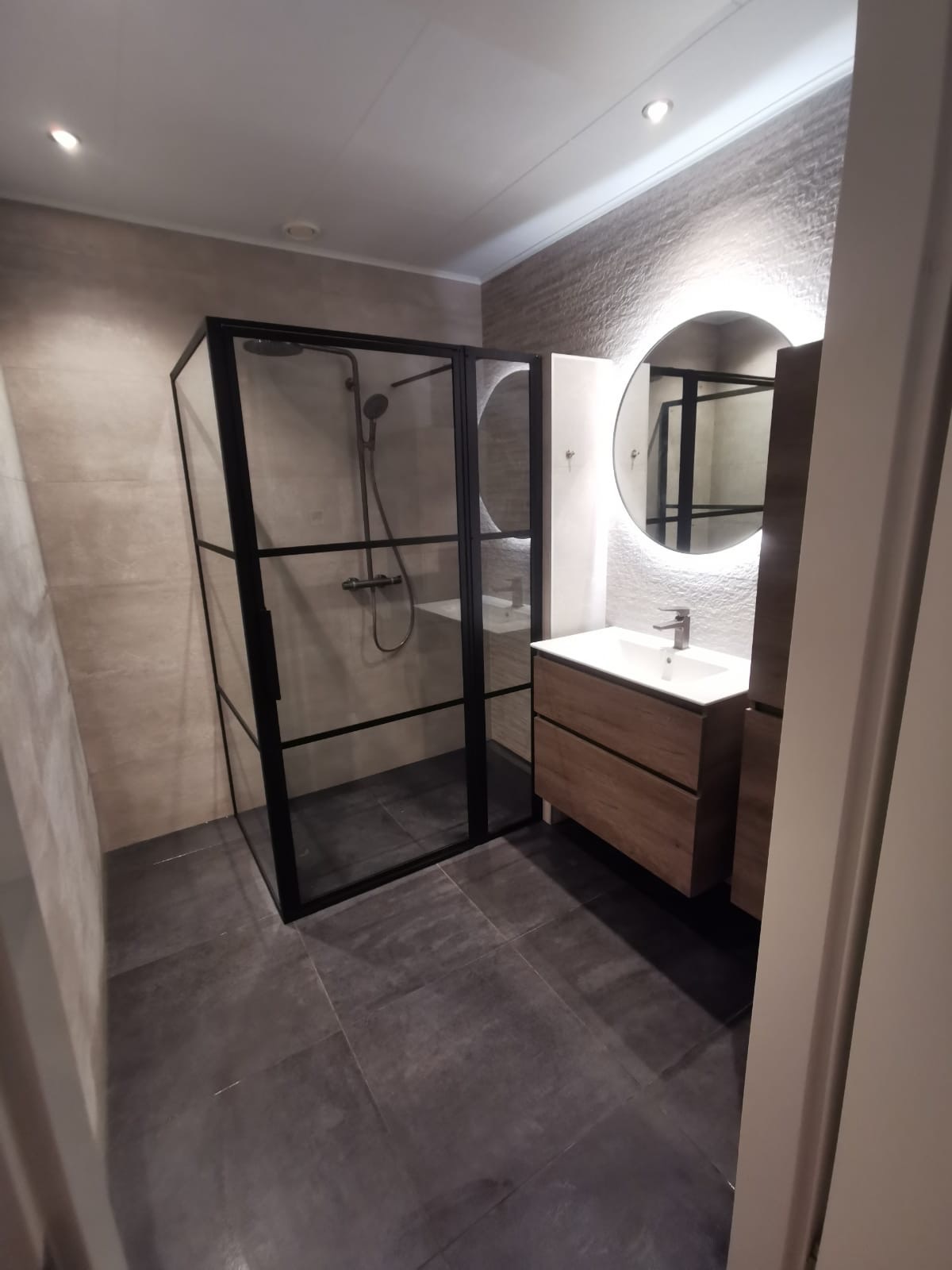 Nowoczesna łazienka z kabiną prysznicową typu walk-in z czarnymi profilami, drewnianą szafką pod umywalką i okrągłym lustrem z podświetleniem LED.