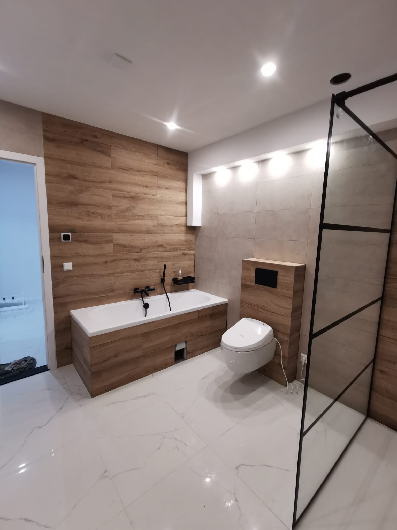 Nowoczesna łazienka z drewnopodobnymi płytkami na ścianach, białą wanną obudowaną drewnem, toaletą myjącą i kabiną prysznicową z czarnymi profilami.