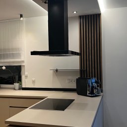 Usługi Remontowe - Nowoczesna kuchnia z beżowymi szafkami, czarną płytą indukcyjną, czarnym okapem, drewnianymi panelami dekoracyjnymi i oświetleniem LED podsufitowym.