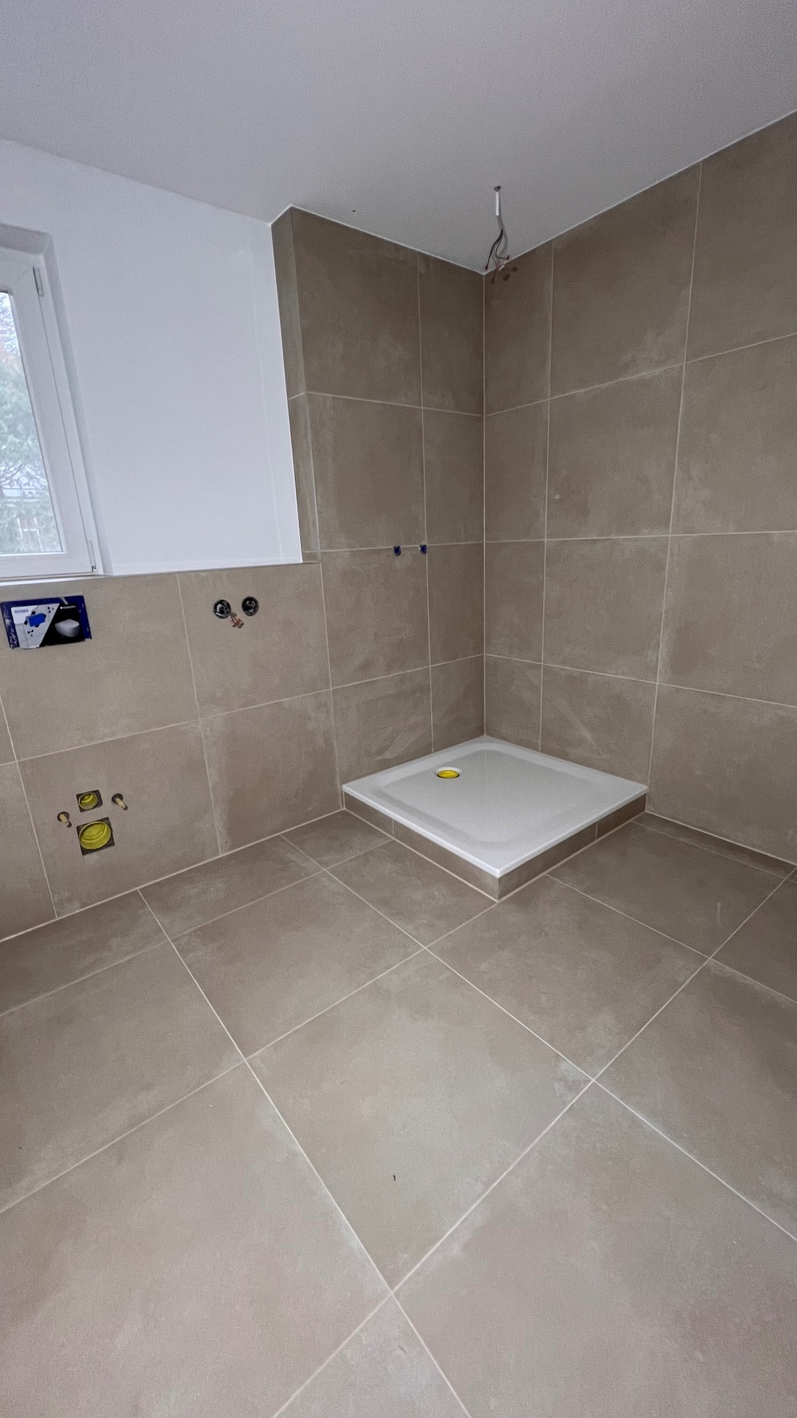 Wnętrze łazienki w trakcie wykańczania, beżowe płytki na ścianach i podłodze, zamontowany biały brodzik, widoczne wyprowadzenia instalacji wodno-kanalizacyjnej.