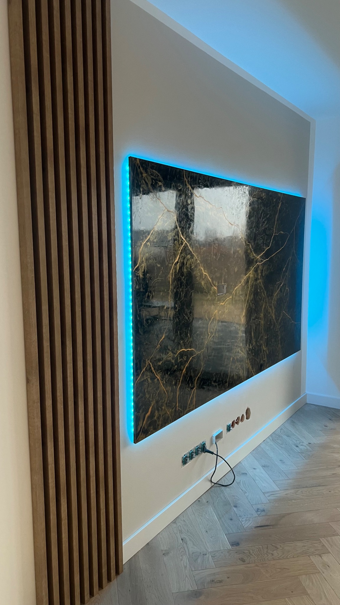 Nowoczesna ściana z dekoracyjnym panelem imitującym marmur, podświetlona niebieskim LED, sąsiadująca z pionowym panelem z drewnianych listewek, widoczne gniazdka elektryczne i parkiet w jodełkę.