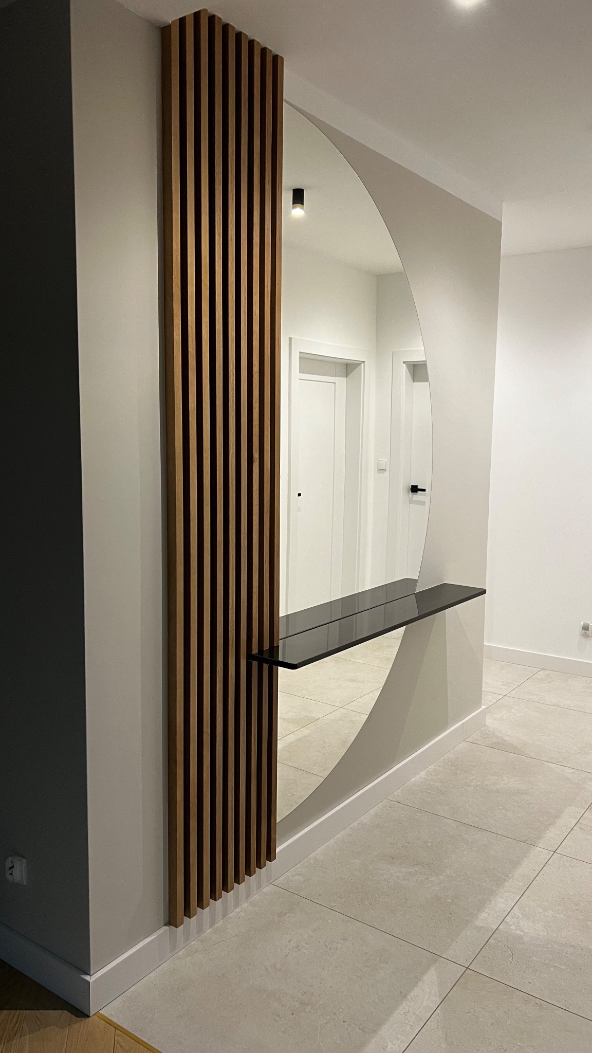 Nowoczesny przedpokój z dekoracyjną ścianką z drewnianych listew, półokrągłym lustrem i czarną, szklaną półką, widok na białe drzwi w tle.