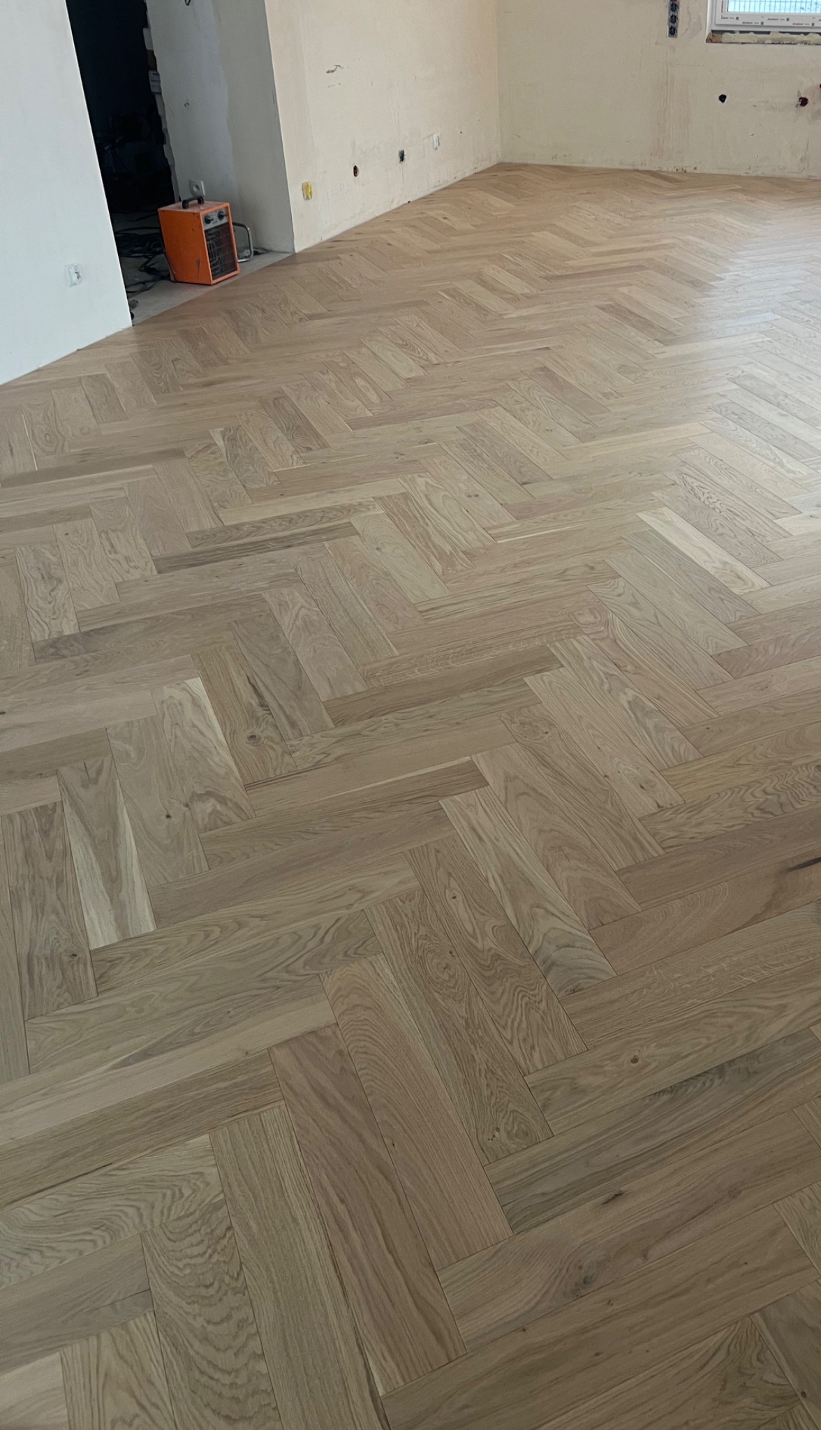 Ułożony parkiet w jodełkę francuską z jasnego drewna w surowym pomieszczeniu z widocznym grzejnikiem elektrycznym i niedokończonymi ścianami.