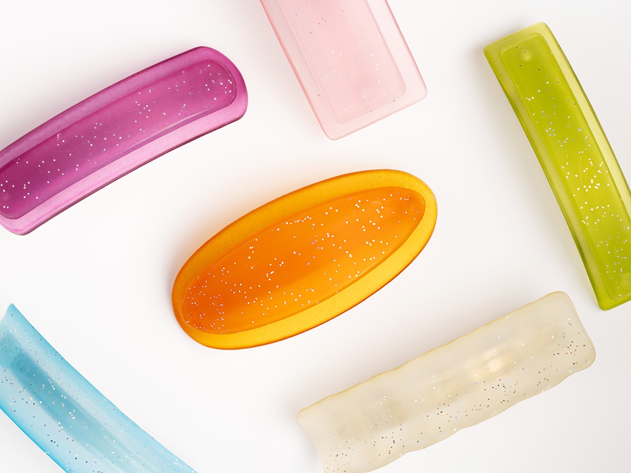 Fotografia produktowa: Różnokolorowe, błyszczące spinki do włosów ułożone na białym tle, widoczne z góry.