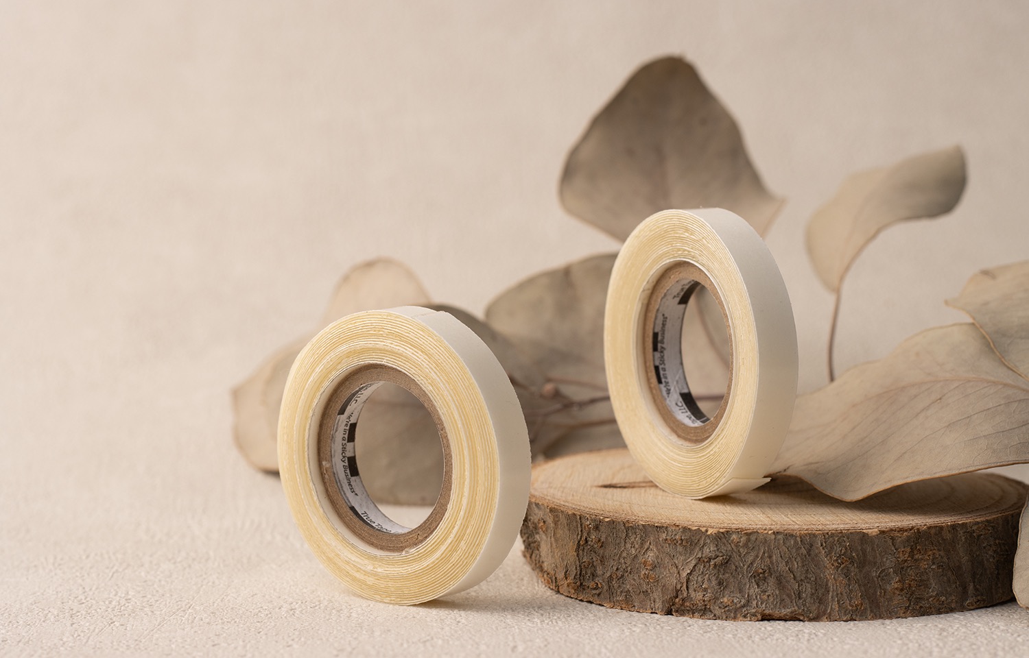 Dwie rolki taśmy dwustronnej na drewnianym krążku, z suszonymi liśćmi eukaliptusa w tle, na beżowej powierzchni.
