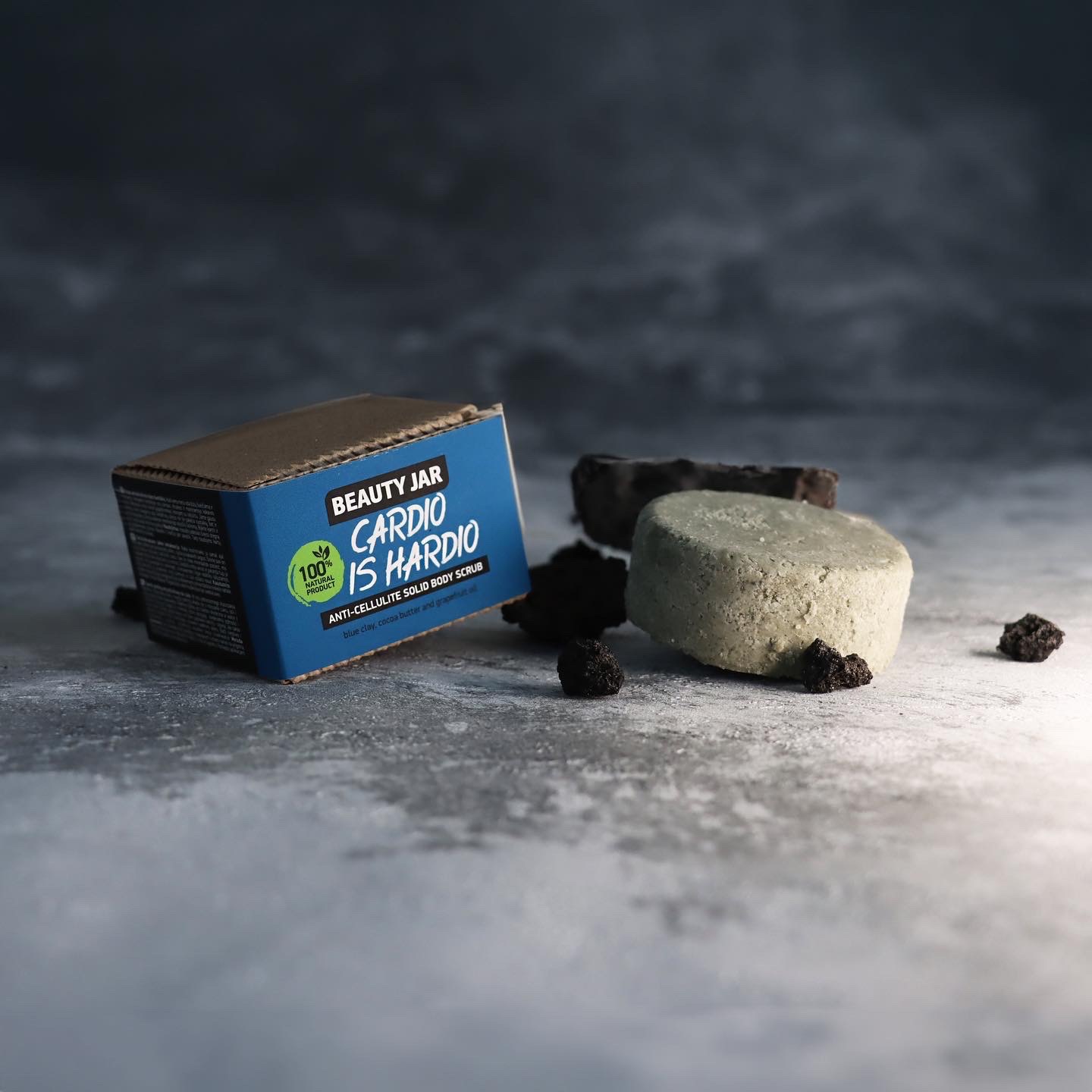 Pudełko antycellulitowego peelingu w kostce Beauty Jar 'Cardio is Hardio' obok dwóch kostek peelingu i okruchów na szarym tle.