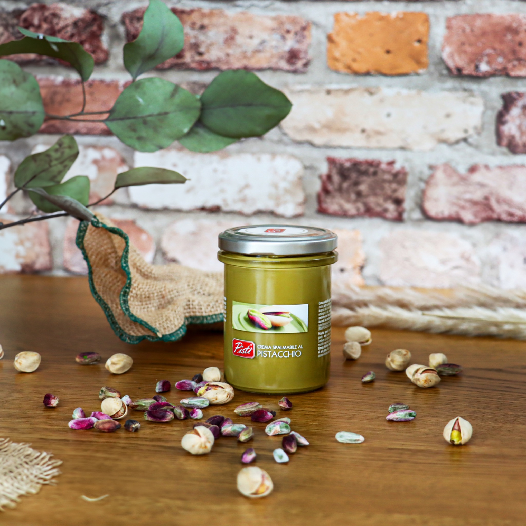 Słoik pistacjowego kremu do smarowania Pisti na drewnianym stole, otoczony rozsypanymi pistacjami, z dekoracyjnym tłem z cegły i zielonymi gałązkami.