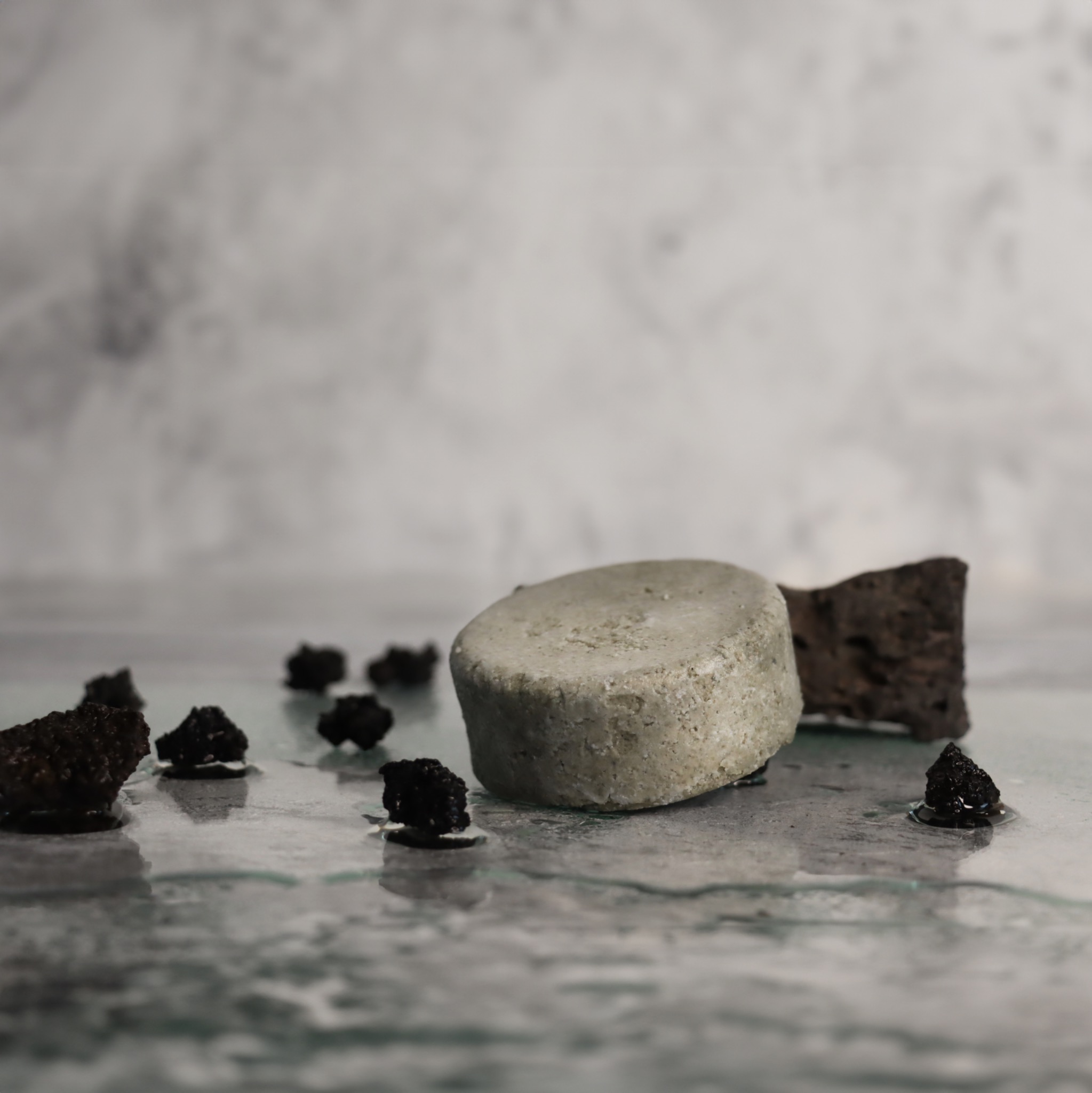 Szare, okrągłe mydło w kostce, obok kawałek pumeksu i kilka czarnych bryłek na mokrej, marmurkowej powierzchni.