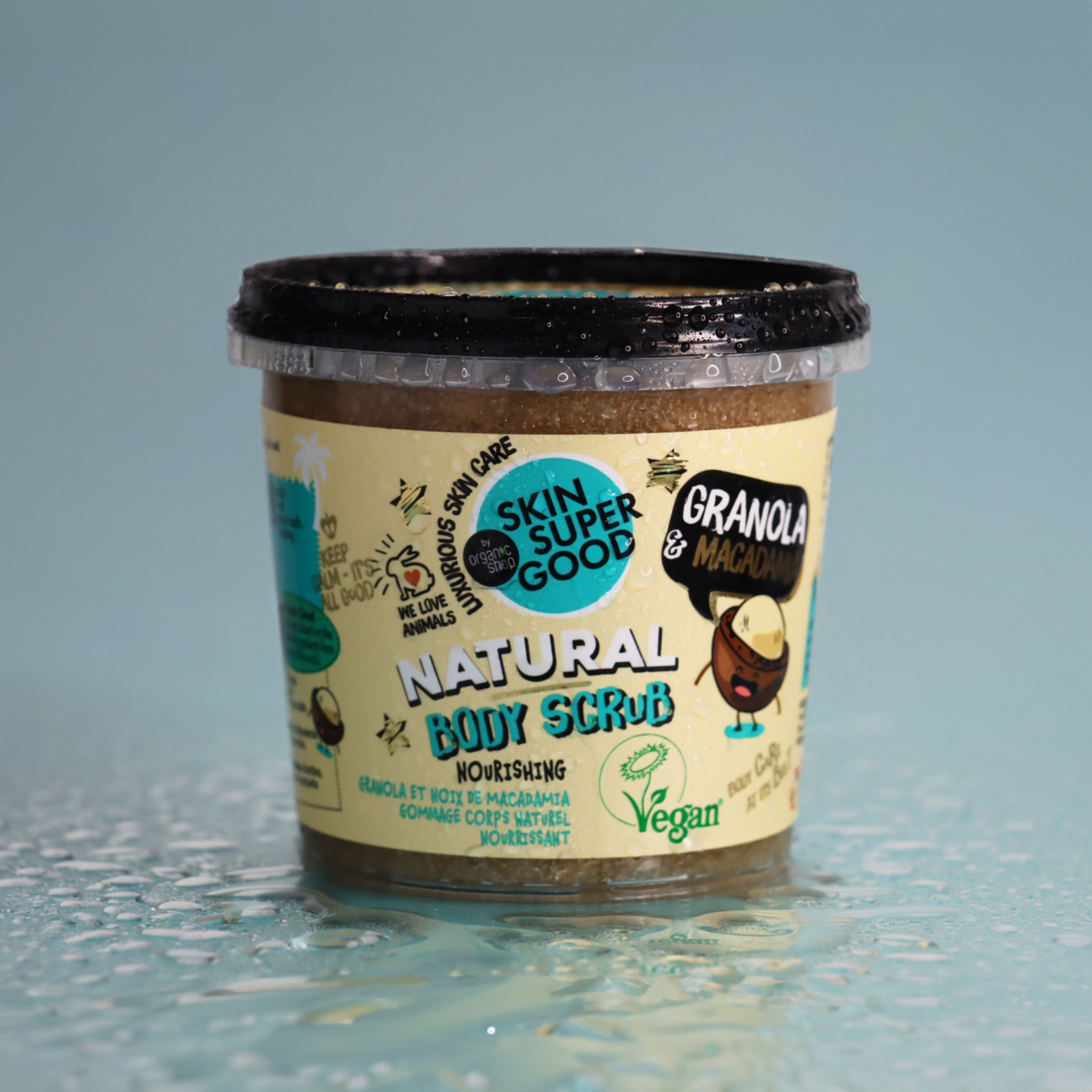 Słoik z naturalnym peelingiem do ciała o zapachu granoli i makadamii, z etykietą w pastelowych kolorach, otoczony kroplami wody na jasnoniebieskim tle.