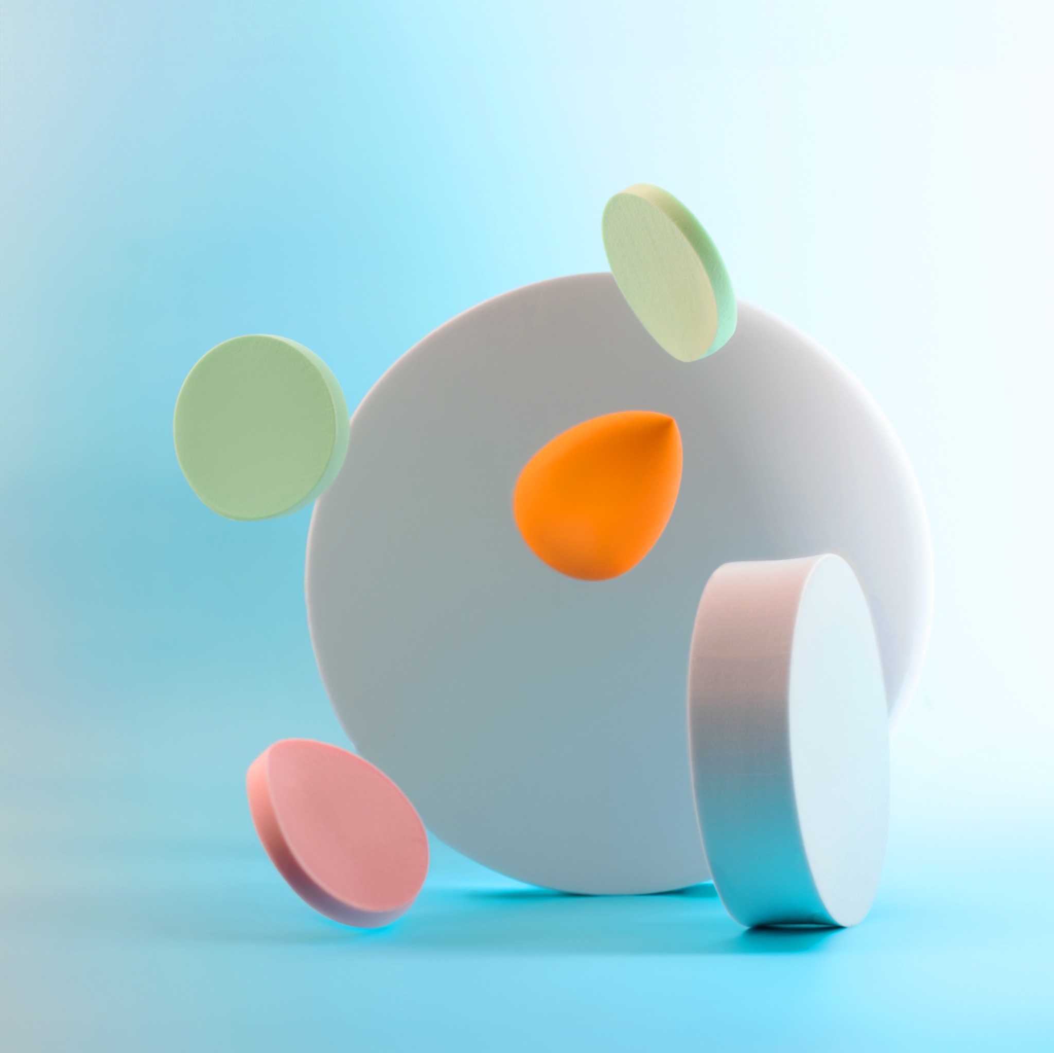 Abstrakcyjna kompozycja z pastelowych, geometrycznych kształtów: okręgi w kolorach zielonym, różowym, biały dysk i pomarańczowa łezka, zawieszone w przestrzeni na błękitnym tle.