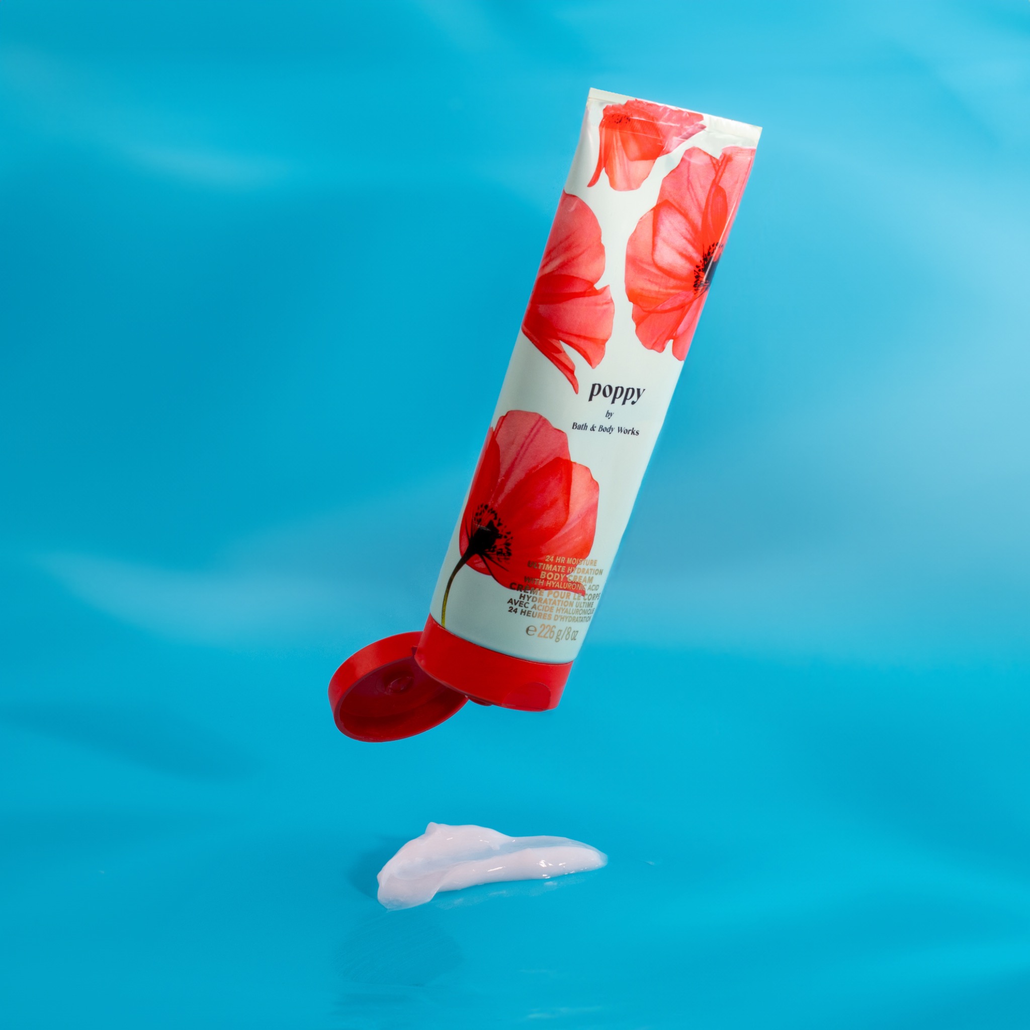 Krem do ciała Poppy marki Bath & Body Works w tubce z czerwonymi makami, z odkręconą czerwoną nakrętką i wyciśniętą porcją kremu na turkusowym tle.