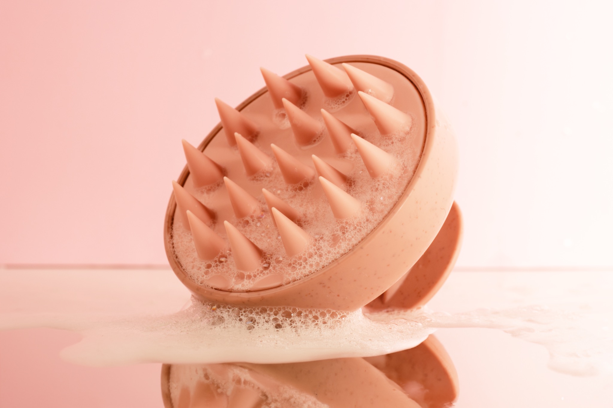 Różowa silikonowa szczotka do masażu skóry głowy z wypustkami, pokryta pianą, odbijająca się w błyszczącym podłożu na tle różowego tła.