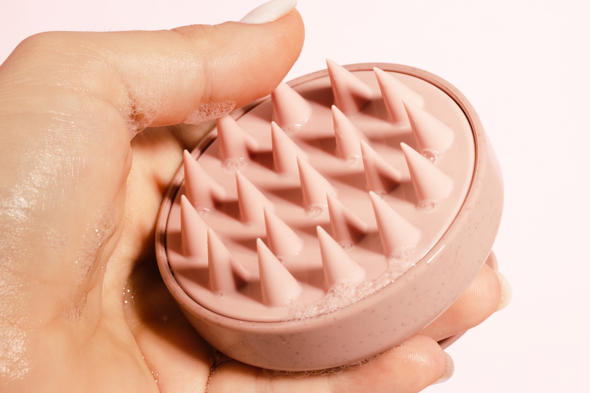 Różowa szczotka do masażu skóry głowy z gumowymi wypustkami, trzymana w dłoni z widoczną pianą na wypustkach, na jasnoróżowym tle.