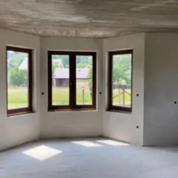 Surowe wnętrze w trakcie wykończenia: ściany pokryte świeżo nałożoną warstwą wygładzającą, trzy okna z brązowymi ramami wpuszczają światło na betonową podłogę, widok na zieleń za oknami.