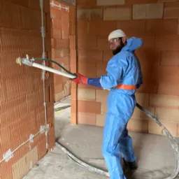 Pracownik w niebieskim kombinezonie i czerwonych rękawicach aplikuje maszynowo tynk na ścianę z cegieł ceramicznych dziurawki, widoczne przewody i złącza elektryczne.