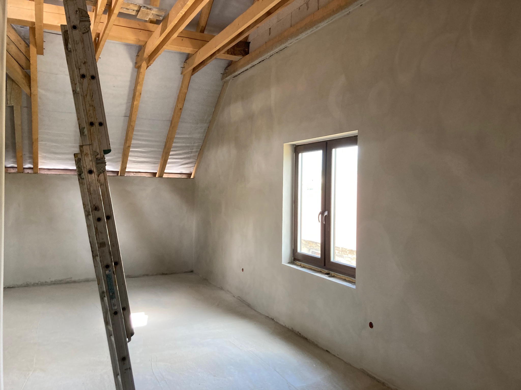 Surowe wnętrze poddasza z oknem i drewnianą konstrukcją dachu, ściany pokryte świeżym tynkiem, oparta drabina.