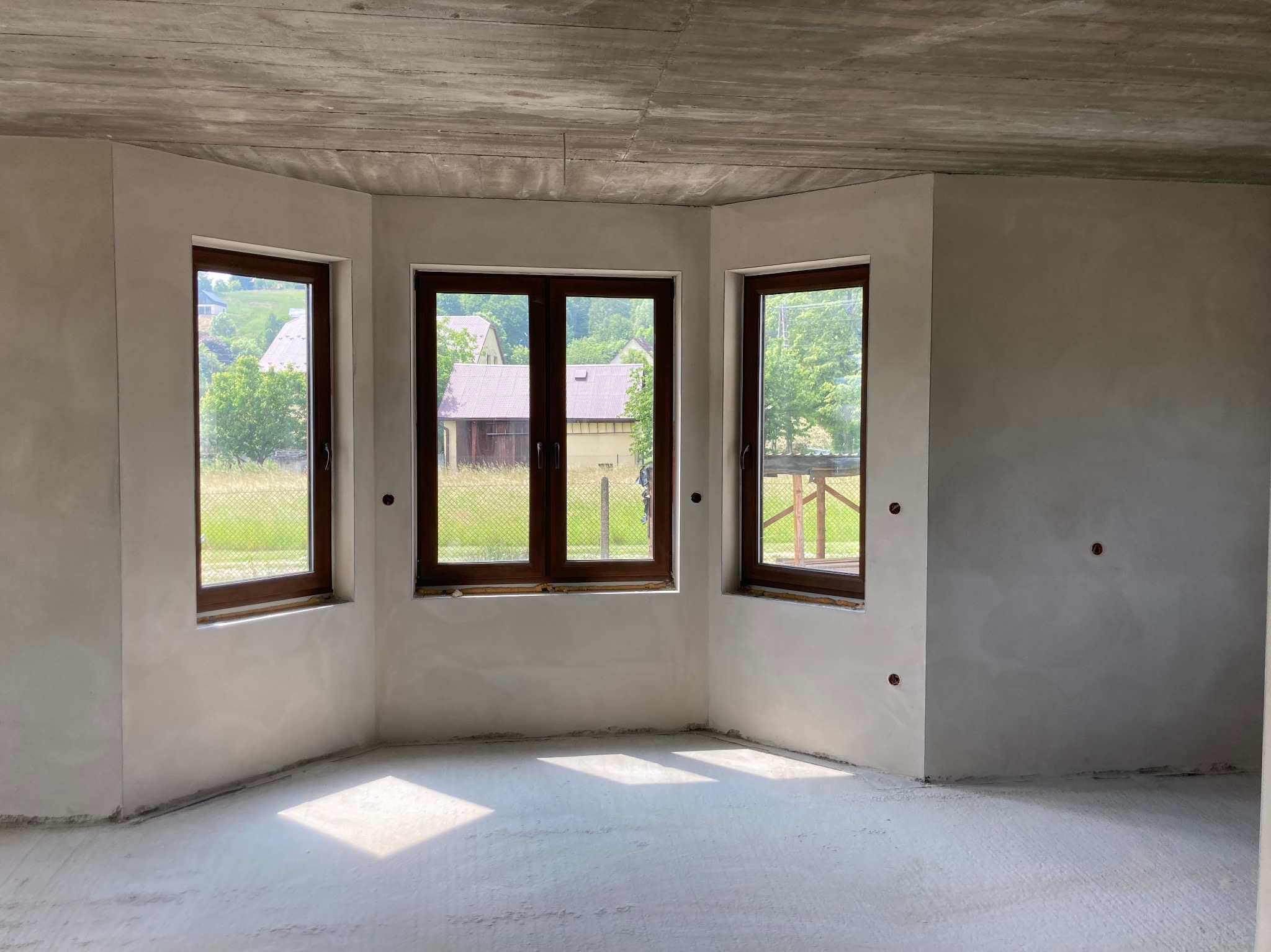 Surowe wnętrze w trakcie wykończenia: ściany pokryte świeżo nałożoną warstwą wygładzającą, trzy okna z brązowymi ramami wpuszczają światło na betonową podłogę, widok na zieleń za oknami.