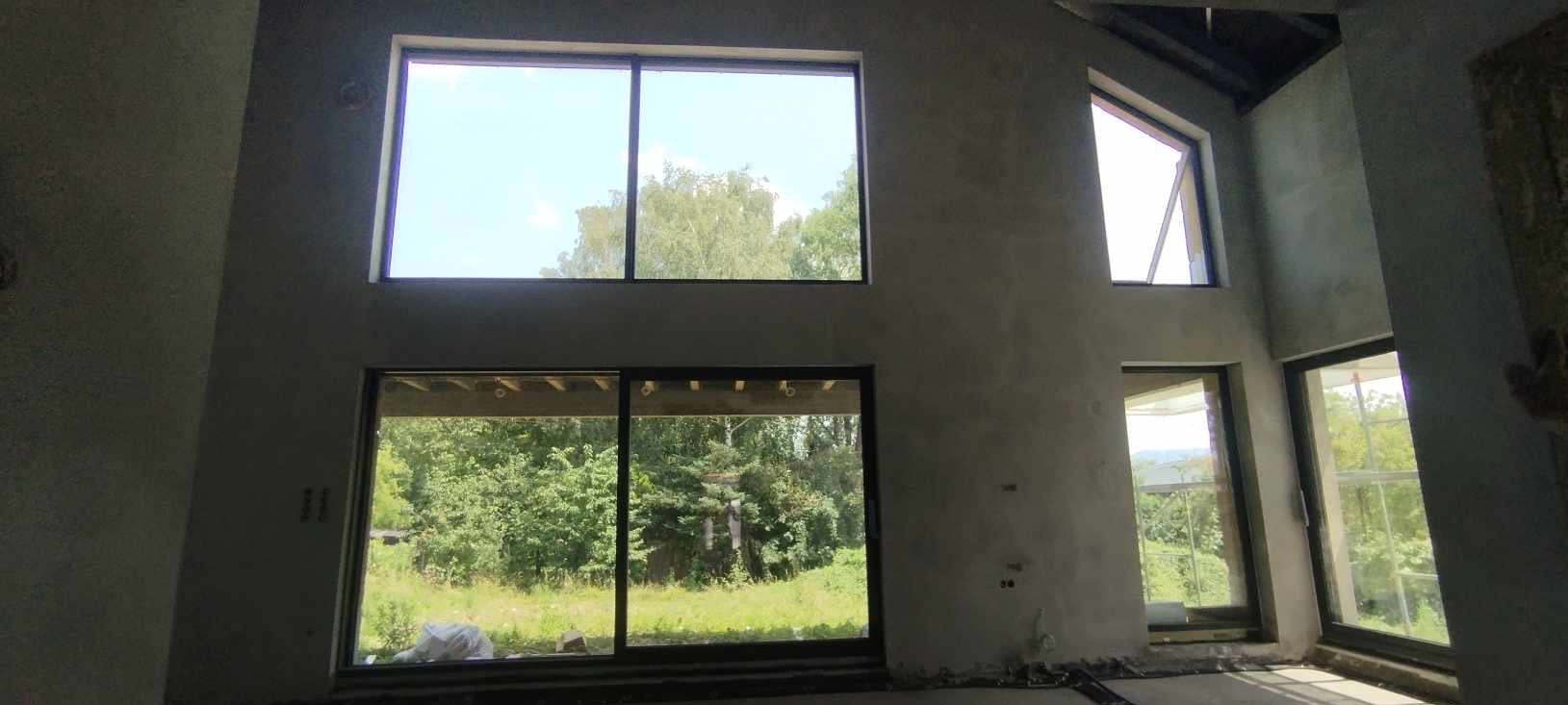 Surowy stan wnętrza z dużymi oknami, ściany pokryte świeżym tynkiem w kolorze szarości, widok na zieleń za oknami, wysoki sufit.