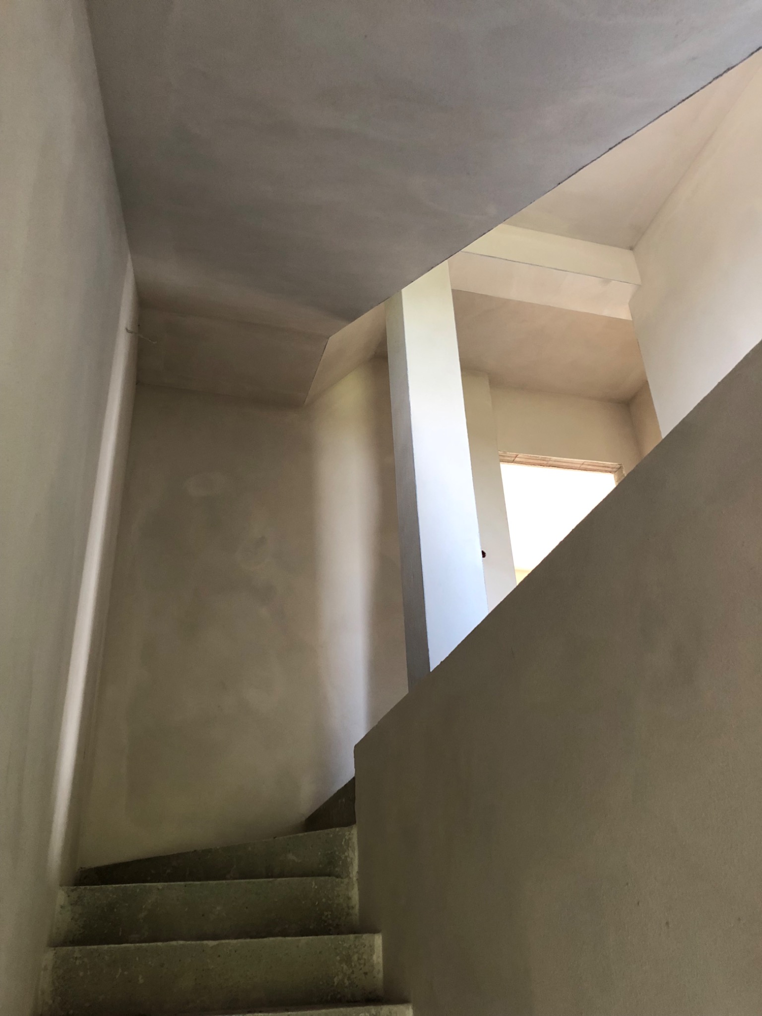Surowe betonowe schody w jasnym wnętrzu w trakcie prac wykończeniowych, ściany i sufit po wstępnym wyrównaniu, widoczna struktura niedoskonałości.