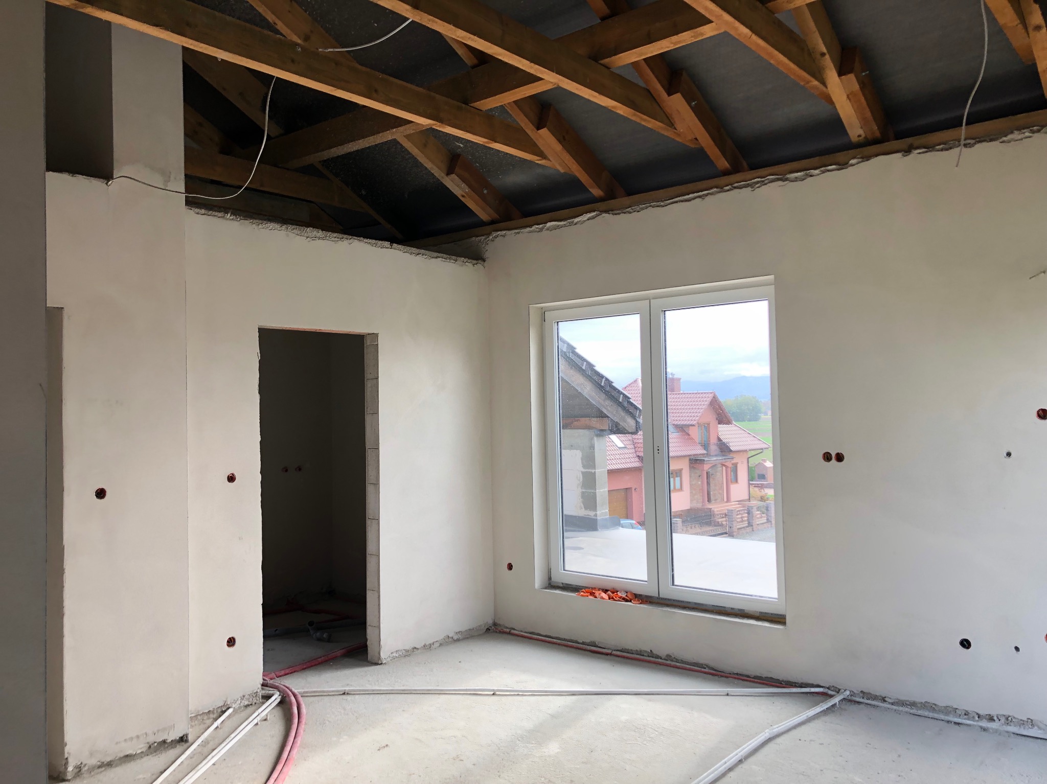Wnętrze w stanie surowym po nałożeniu pierwszej warstwy gładzi, widoczne drewniane elementy konstrukcji dachu, instalacje elektryczne w ścianach i rurki na podłodze, okno z widokiem na dom.