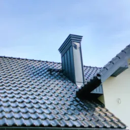 Błyszcząca, grafitowa dachówka ceramiczna z kominem w nowoczesnym stylu, rynna w kolorze dachu i kamera przemysłowa pod okapem na tle błękitnego nieba.