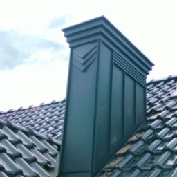 Metalowy komin z dekoracyjnymi elementami na błyszczącym, ciemnym dachu z dachówki ceramicznej pod pochmurnym niebem.