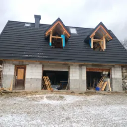 Dom w trakcie budowy z zamontowaną, czarną dachówką i oknami dachowymi w trakcie montażu, widoczna więźba dachowa z zabezpieczeniem przeciwwilgociowym.