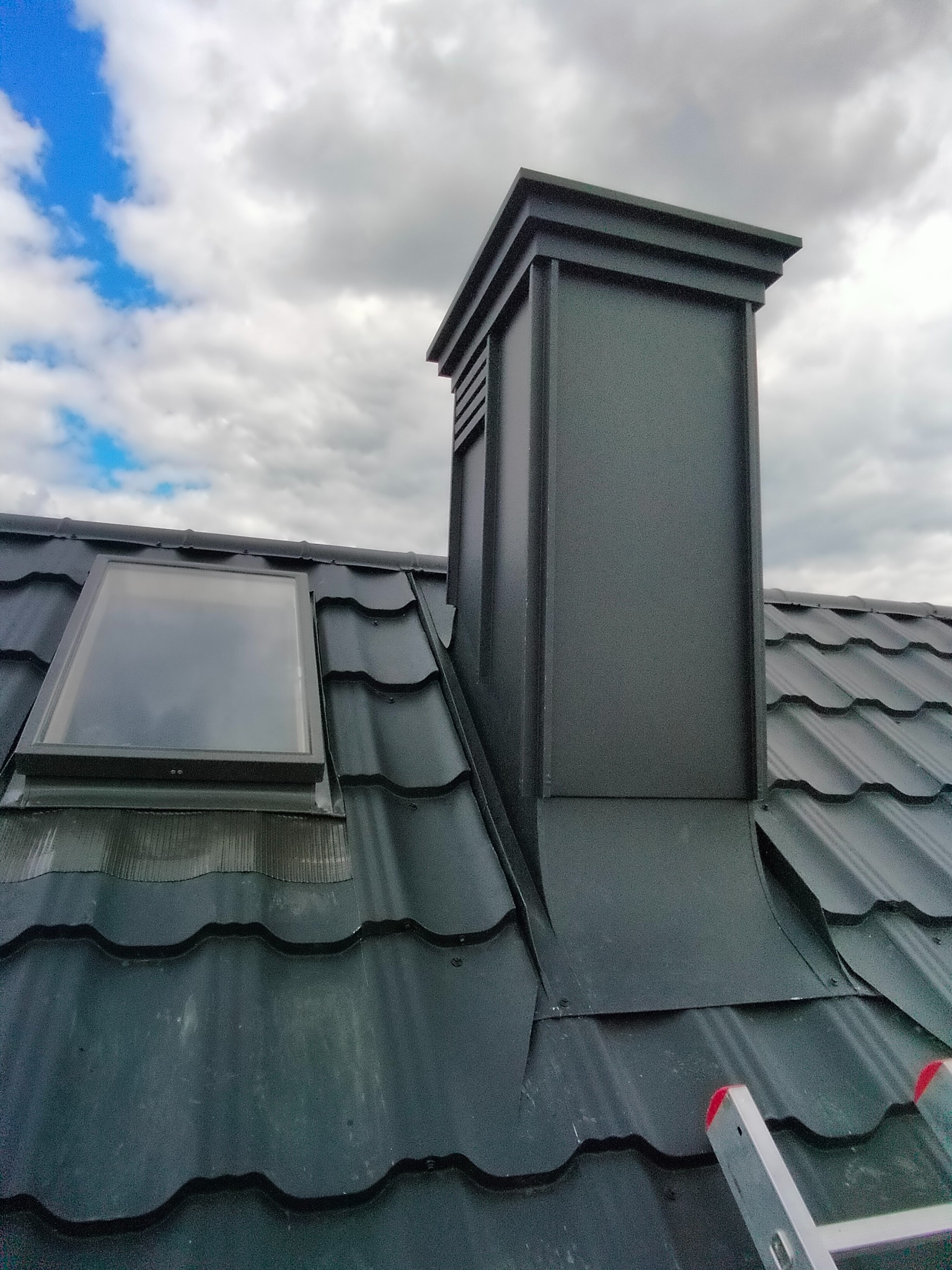 Czarny dach z blachy trapezowej z oknem dachowym i kominem wentylacyjnym na tle pochmurnego nieba, widoczny fragment drabiny.