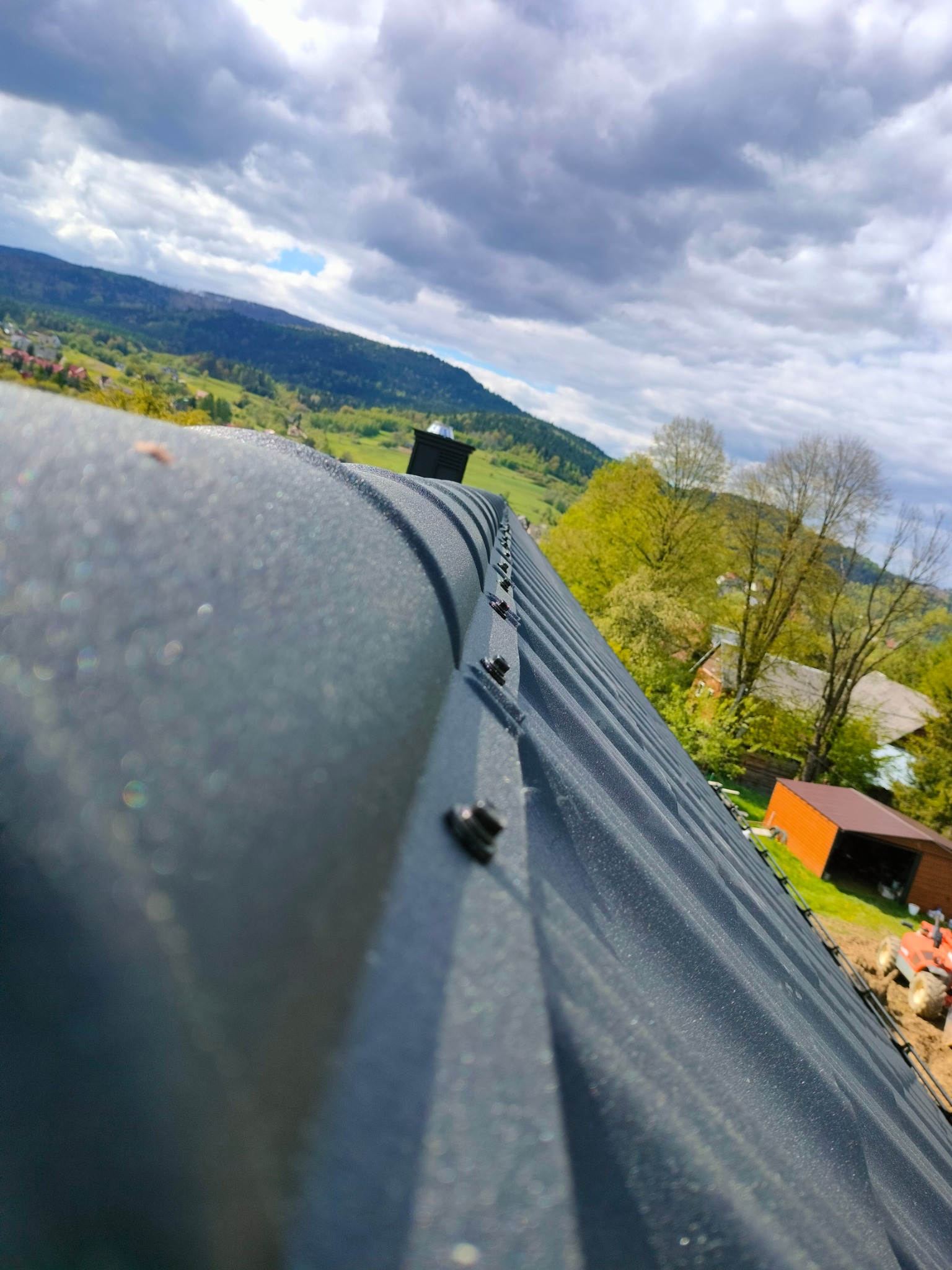 Nowy, grafitowy dach z blachy na tle malowniczego, górzystego krajobrazu i błękitnego nieba z chmurami. Widoczne elementy mocujące i komin.
