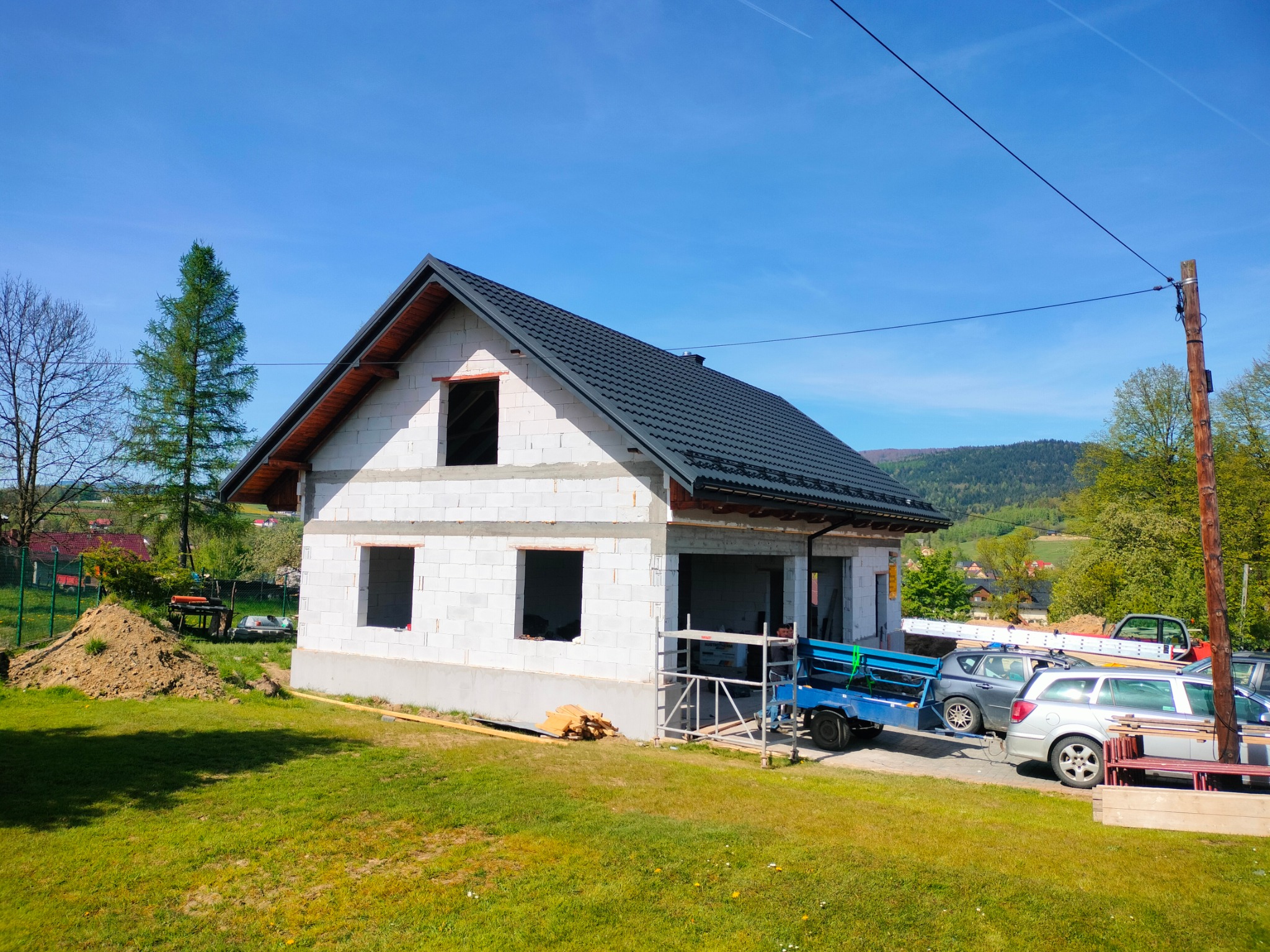 Dom w trakcie budowy z ciemnoszarą dachówką, widoczne rusztowania i przyczepka z materiałami budowlanymi na tle zielonego krajobrazu.