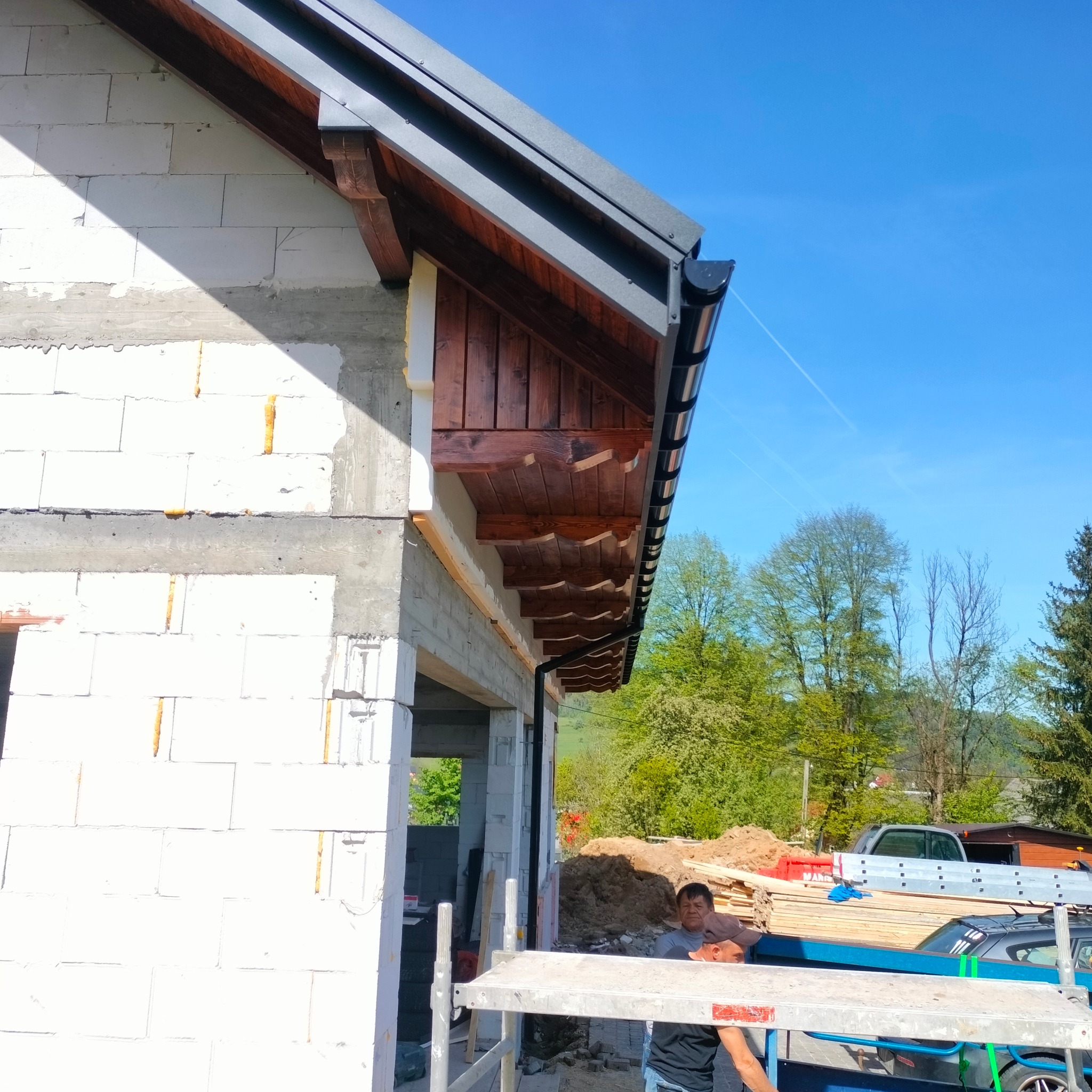 Fragment nowo budowanego domu z widoczną drewnianą konstrukcją dachu, metalowymi rynnami i częściowo wykończoną elewacją z białych bloczków.