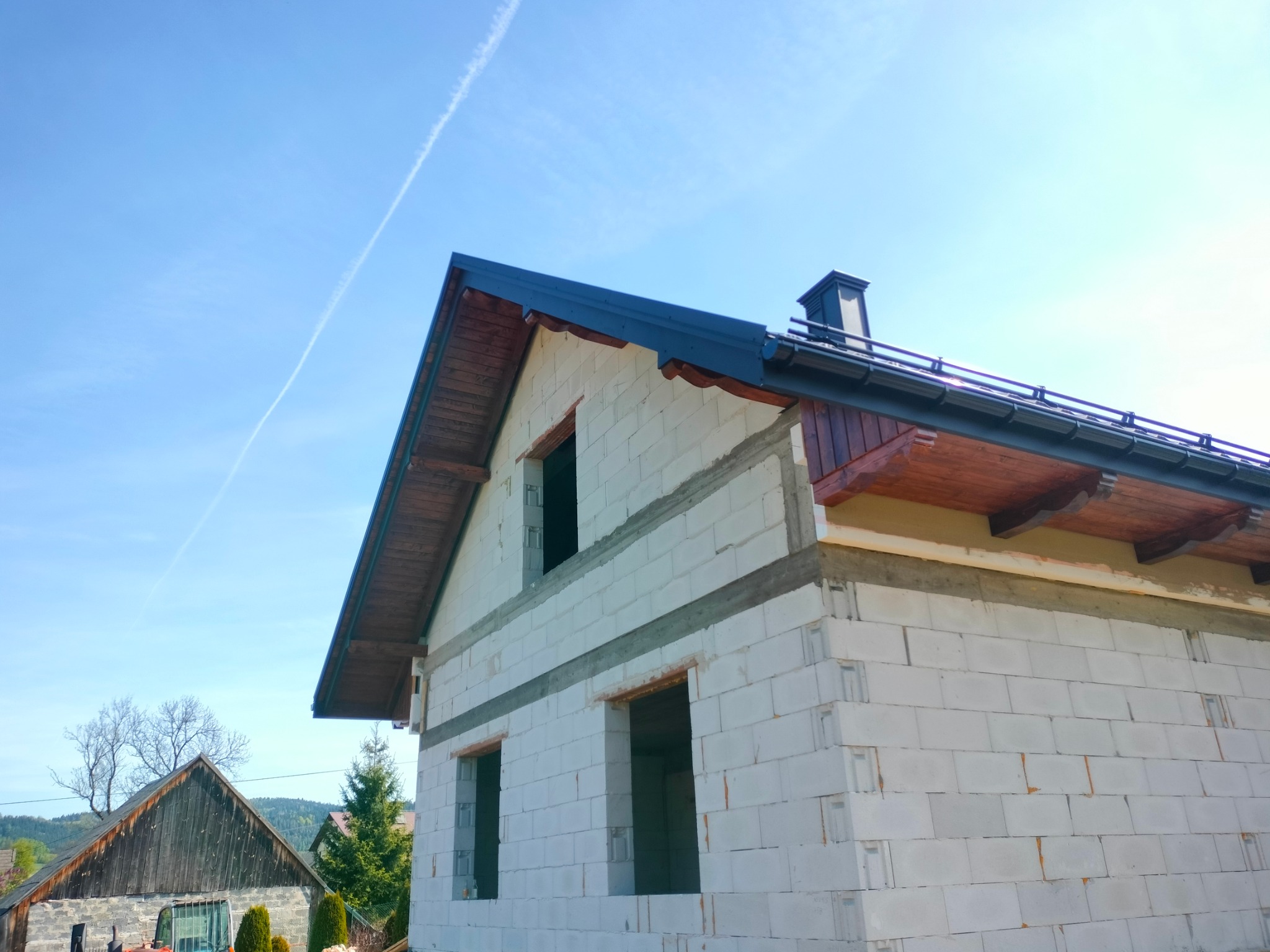 Surowy budynek w stanie deweloperskim z ciemnoszarą blachą na dachu i kominem, widok z dołu na tle błękitnego nieba z smugą kondensacyjną.