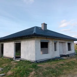 Niedokończony dom jednorodzinny z ciemną, nową dachówką ceramiczną i szarym kominem, widoczne niezabudowane otwory okienne i drzwiowe, teren wokół domu niezagospodarowany.