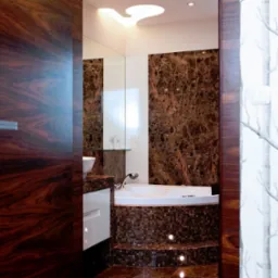 Nowoczesna łazienka z wanną obudowaną mozaiką w odcieniach brązu, marmurową ścianą i fototapetą z motywem drzew, widoczna przez uchylone, ciemne, drewniane drzwi.